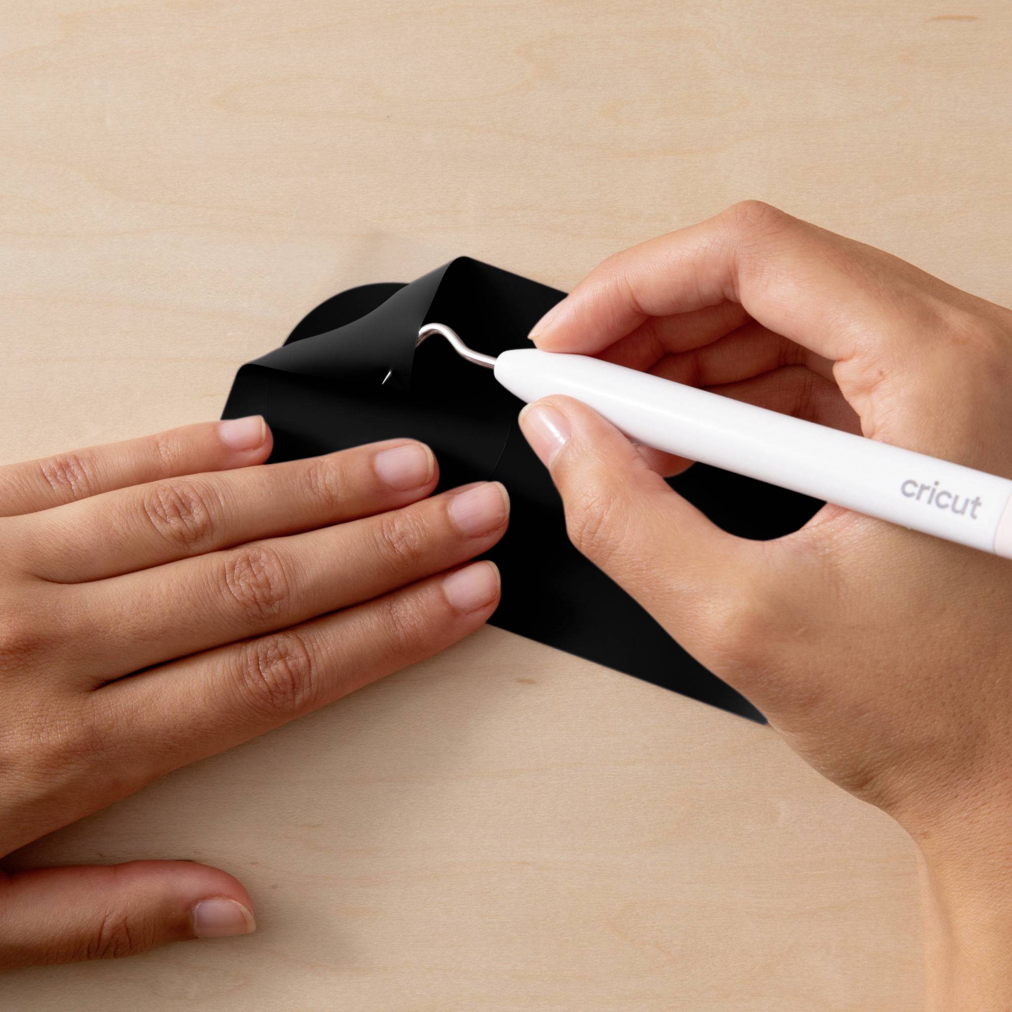 Gros plan sur deux mains. L'une tient un matériau noir, l'autre un outil Cricut pour couper ou travailler.