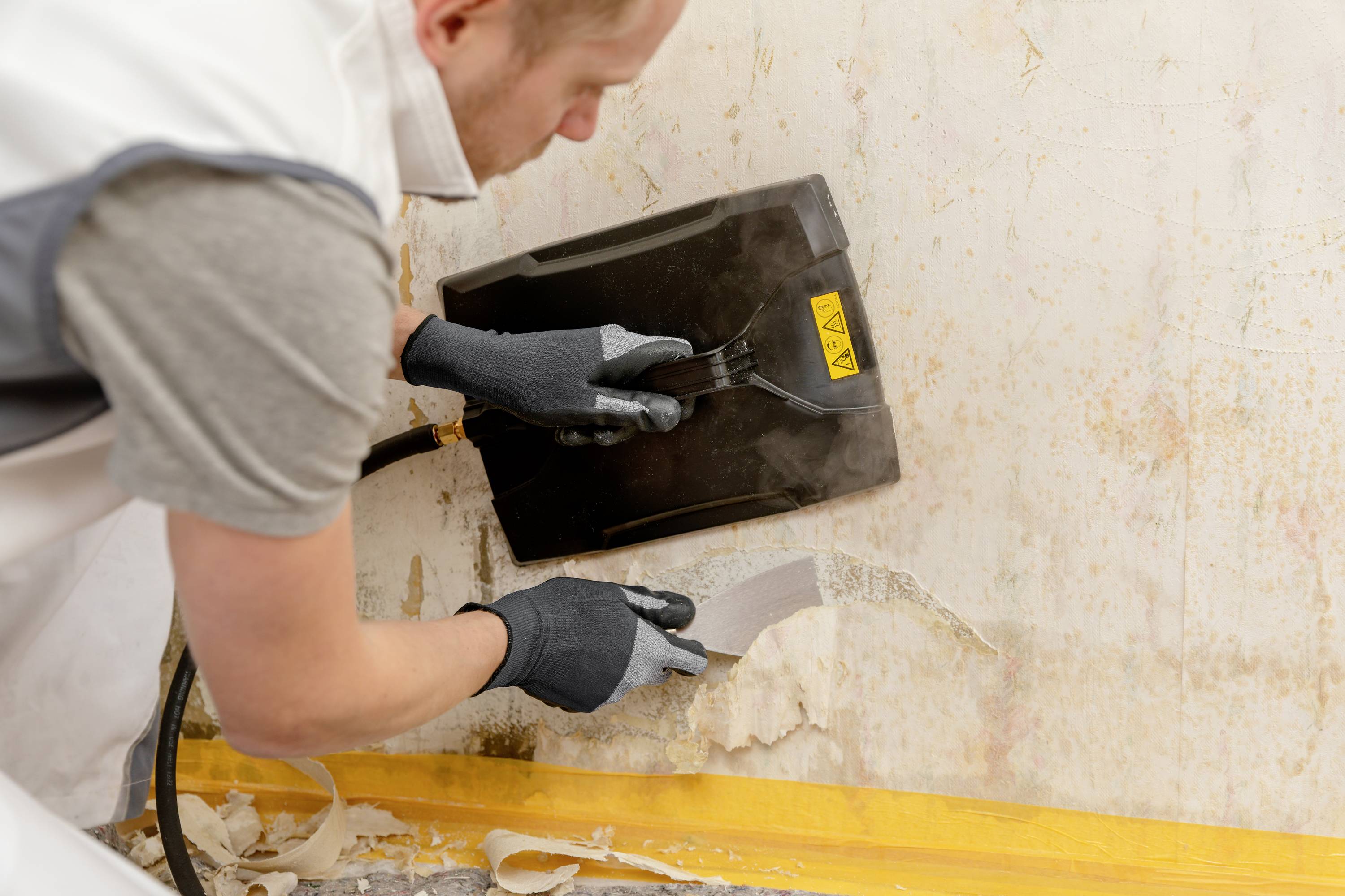Un homme retire du papier peint d'un mur à l'aide d'un décolleur de papier peint à vapeur. Il porte des gants de travail et se concentre sur sa tâche.