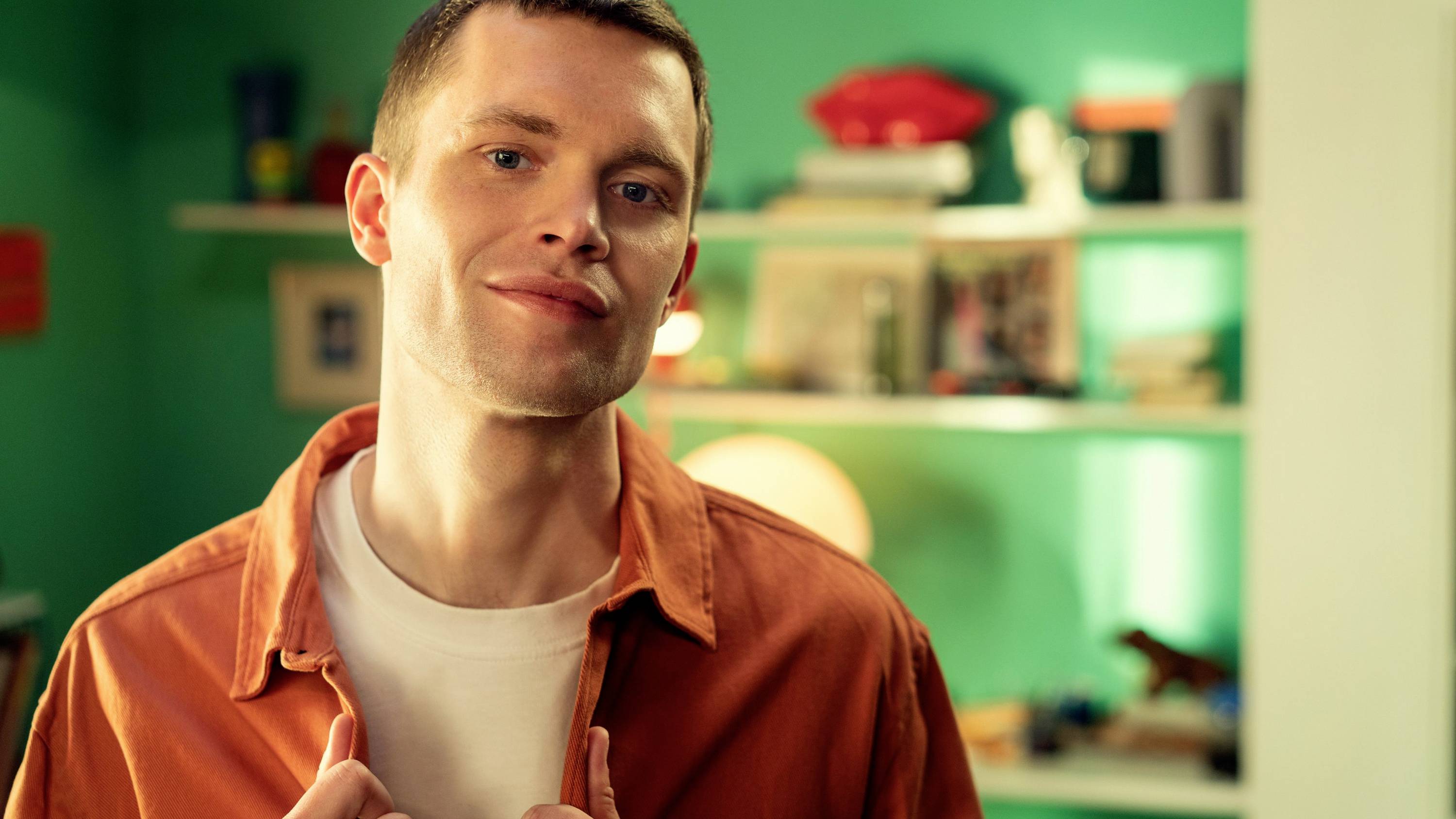 Une personne portant une chemise orange se tient souriante dans une pièce avec un mur vert et des étagères remplies de livres et de décorations.