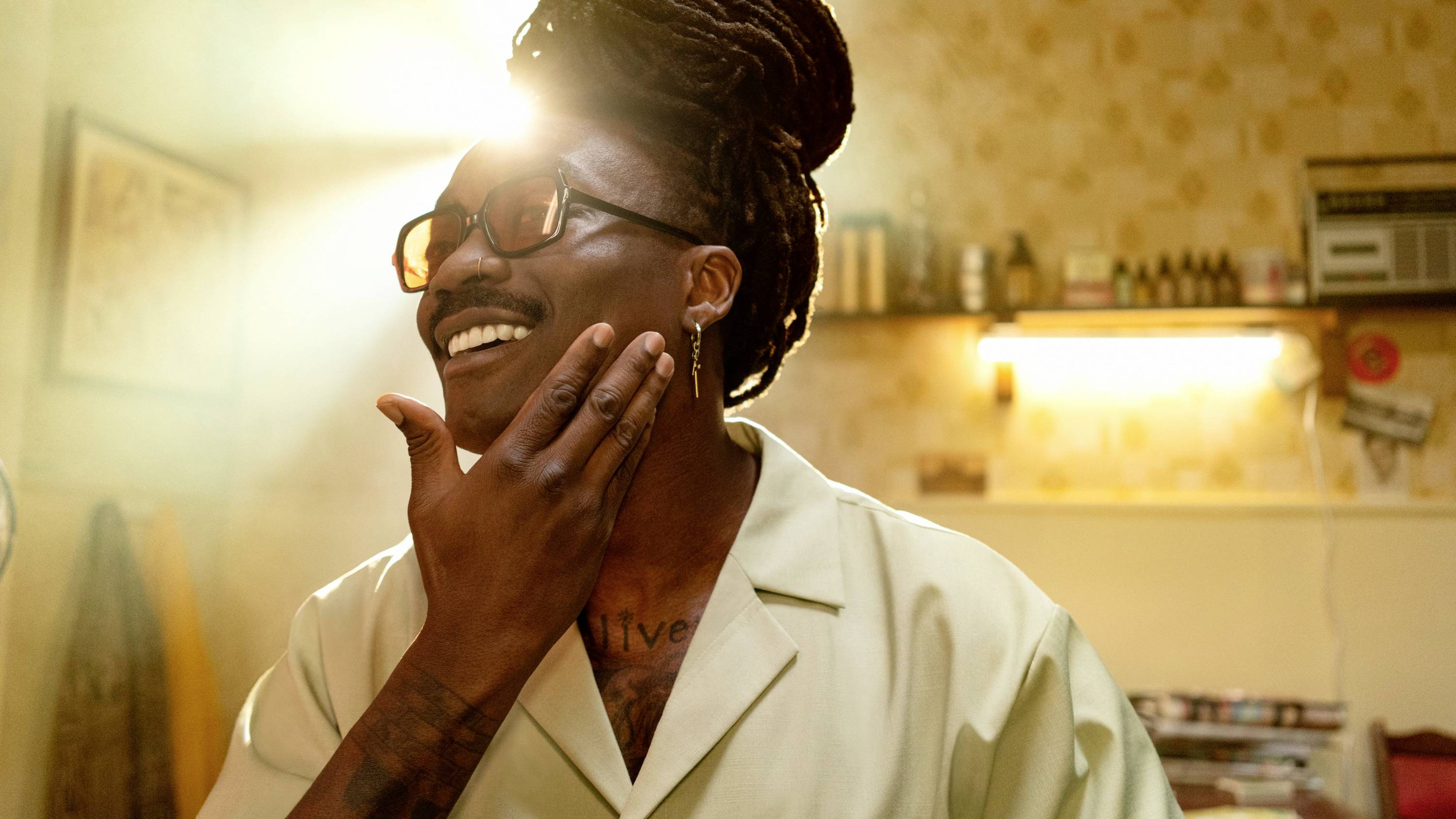 Une personne aux dreadlocks et lunettes se tient souriante dans une pièce, touchant son menton, baignée de lumière. Des étagères sont visibles en arrière-plan.