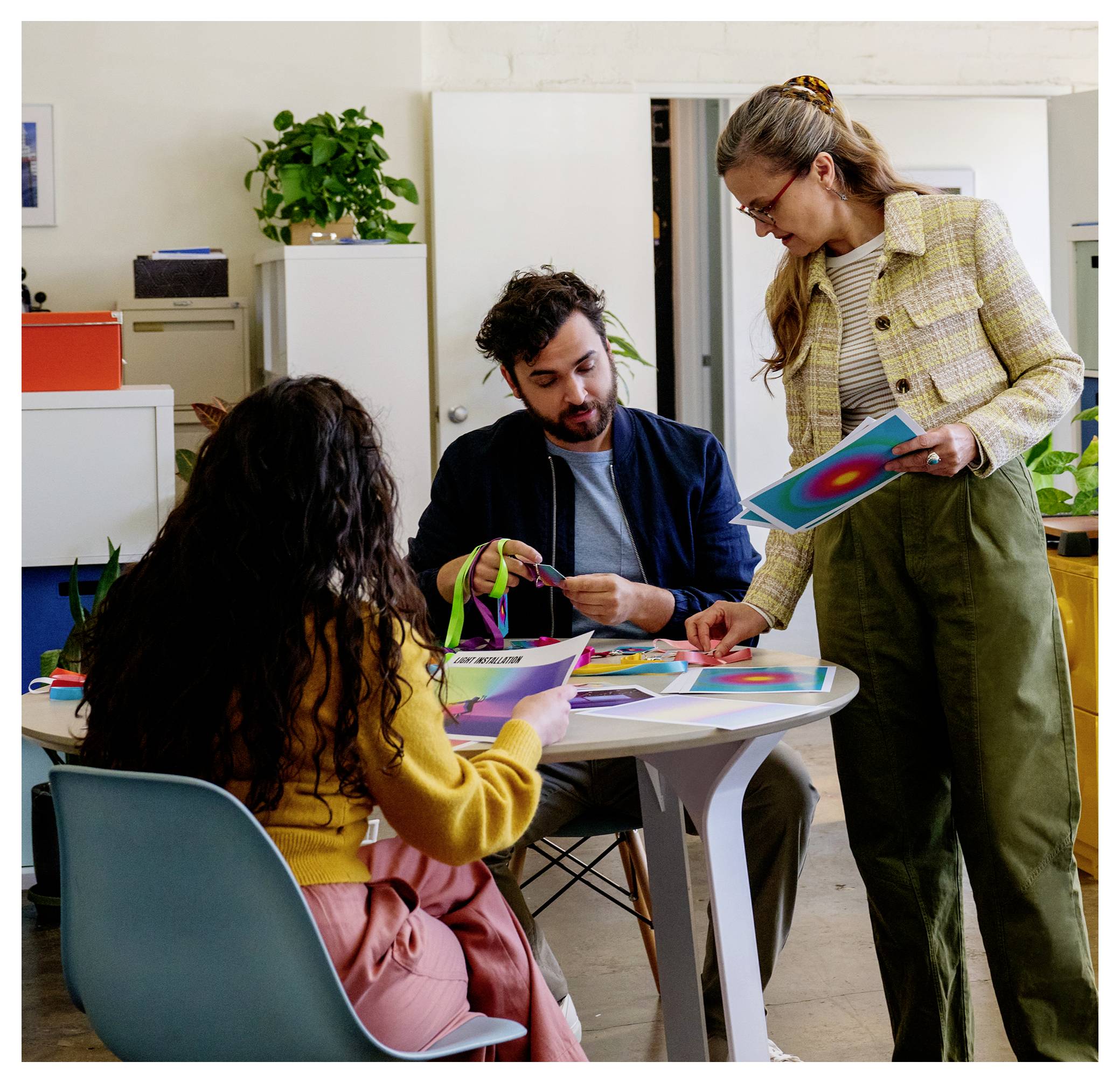 Trois collègues examinent des designs imprimés colorés autour d'une petite table dans un bureau lumineux, une femme debout pointant du doigt tandis que les autres examinent les matériaux.