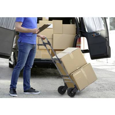 Une personne vêtue de bleu utilise un chariot manuel pour décharger des cartons d'un camionnette ouverte et note quelque chose sur un presse-papiers.