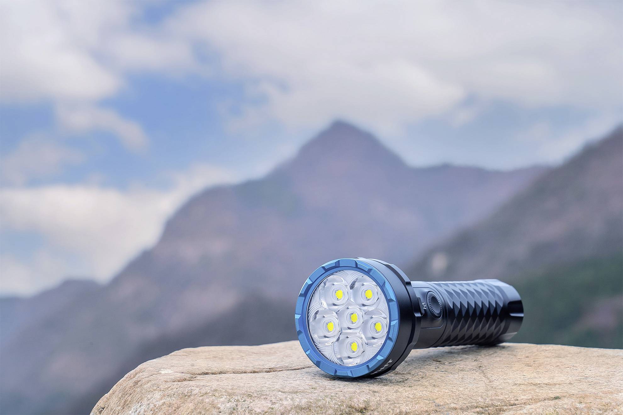 Lampe de poche avec plusieurs LED posée sur un rocher, derrière un paysage montagneux sous un ciel nuageux.