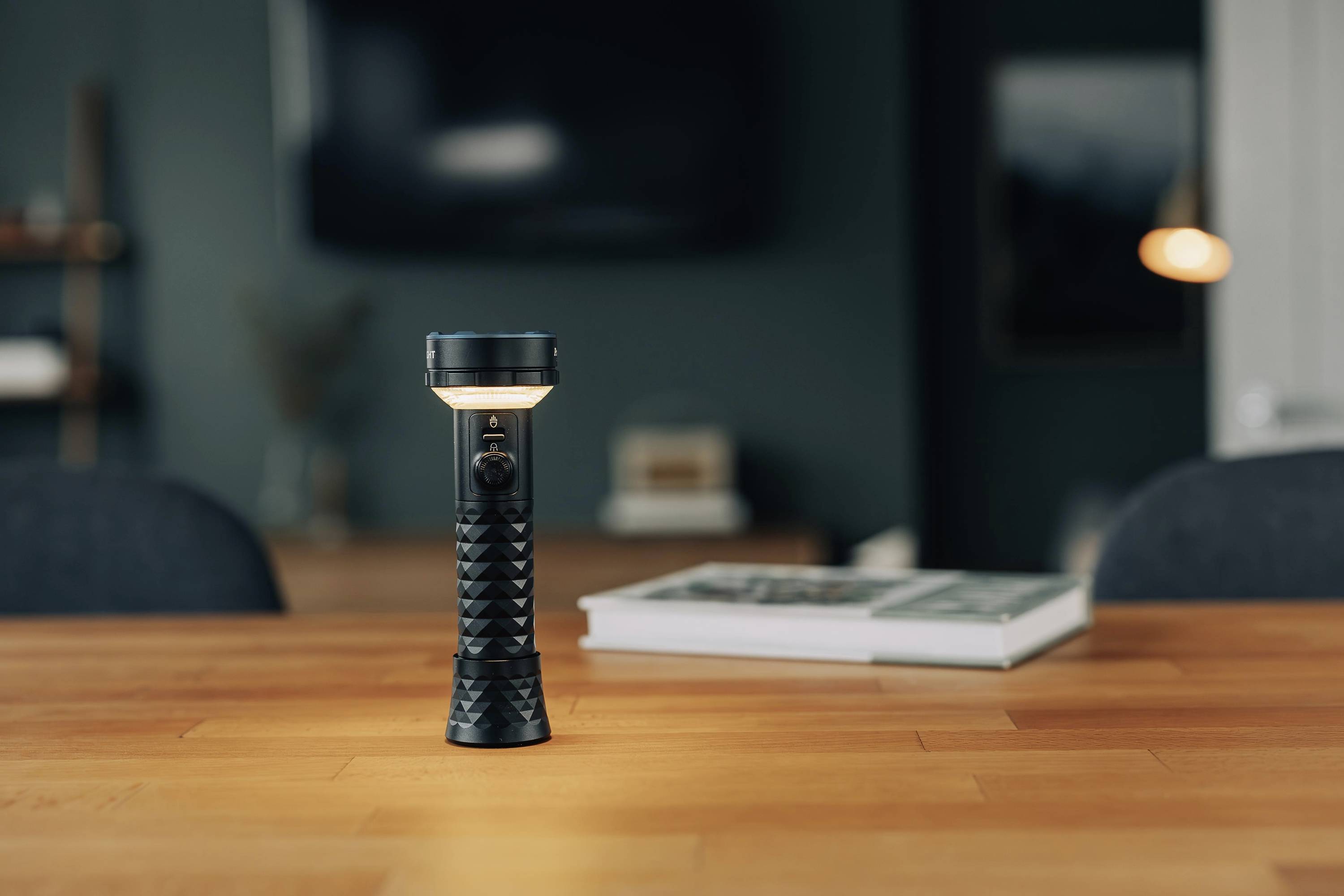 Une lampe de poche allumée est posée sur une table en bois dans une pièce au design moderne, avec des meubles flous visibles en arrière-plan.