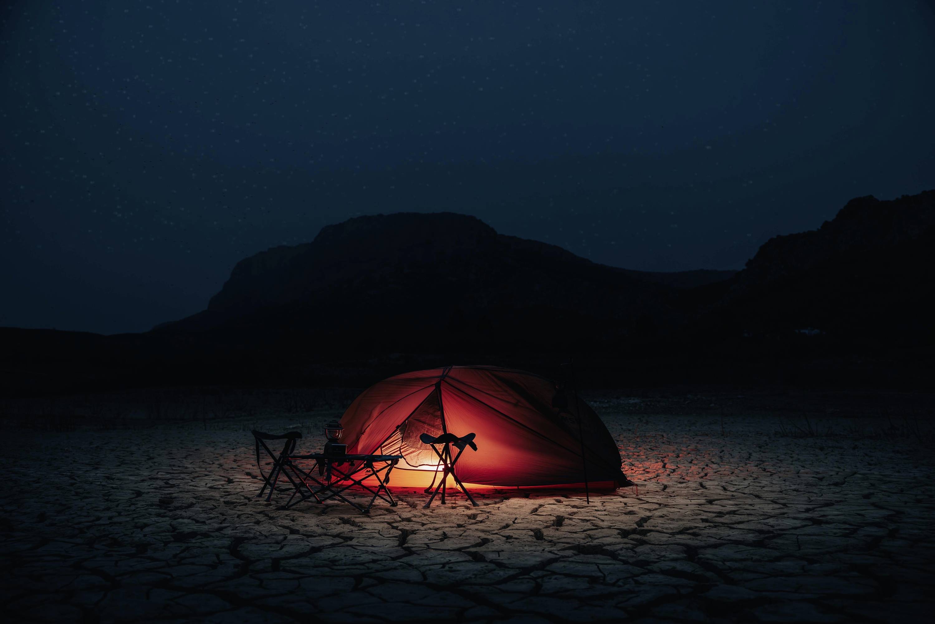 Une tente rouge illuminée et deux chaises de camping vides sont installées sur un sol sec et craquelé, devant une silhouette sombre de montagne, dans la nuit.