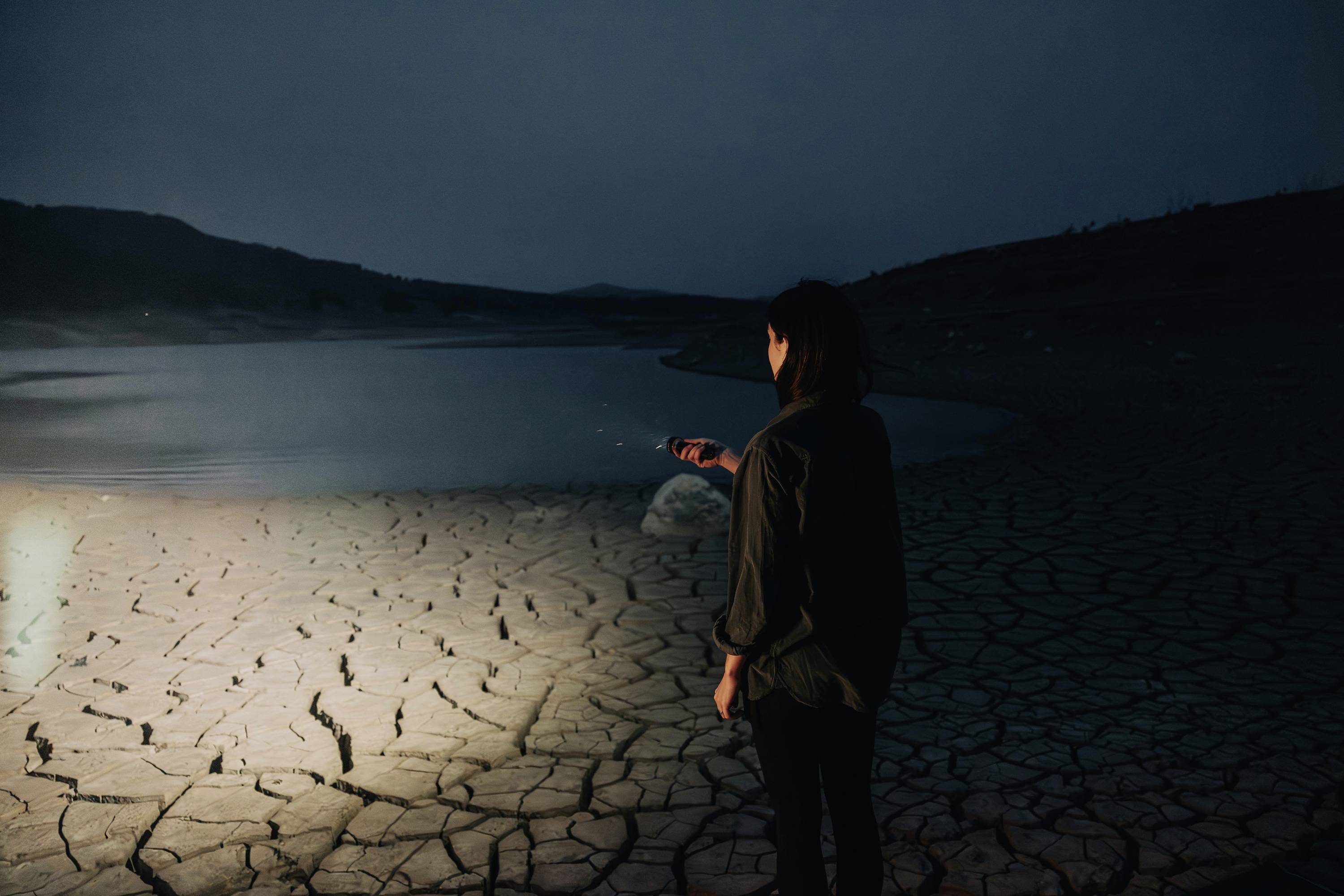 Une personne se tient debout sur un sol sec et craquelé, regardant un petit plan d'eau, tandis que le ciel est sombre.
