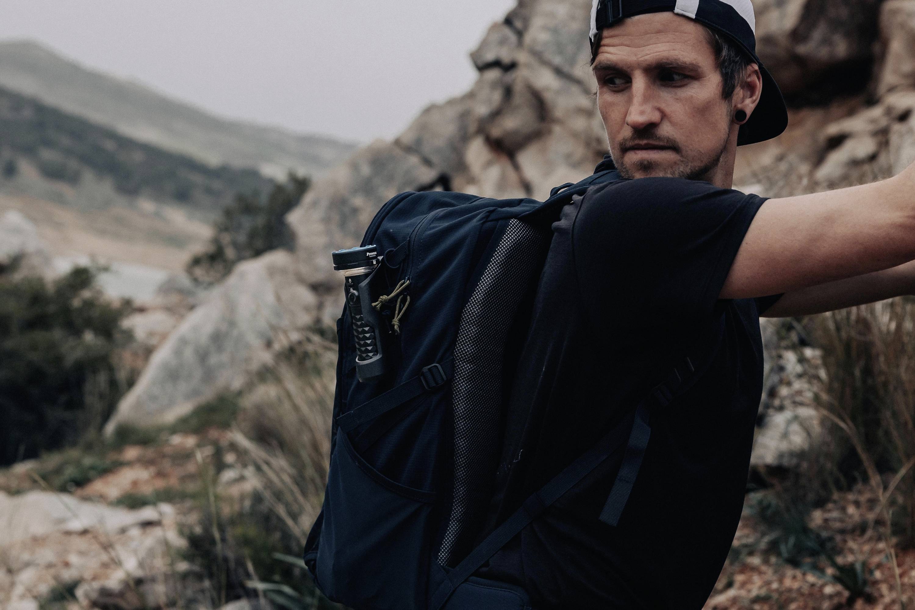 Une personne portant un sac à dos noir marche dans un paysage montagneux. Des rochers et de la végétation sont visibles en arrière-plan.