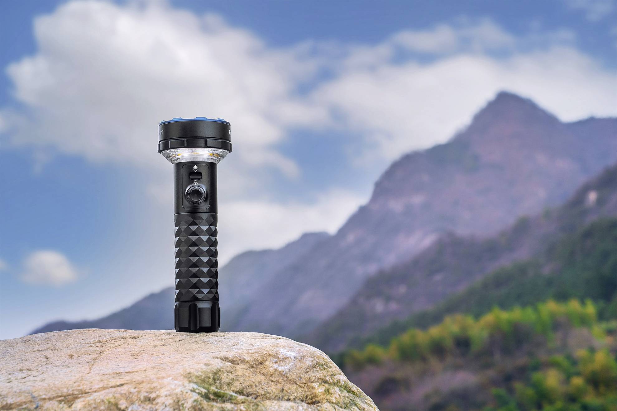 Une lampe de poche noire est posée sur un rocher au premier plan. À l'arrière-plan, on distingue des montagnes floues et un ciel bleu.