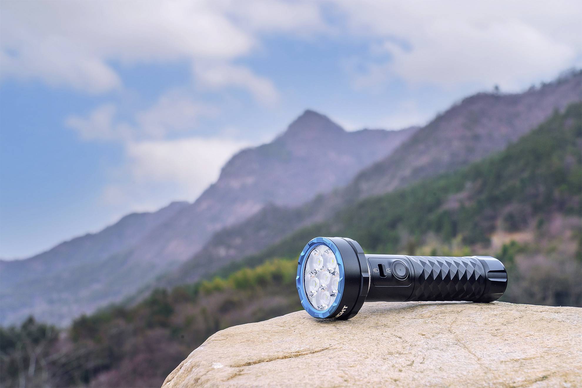 Une lampe de poche est posée sur un rocher au premier plan, avec un paysage montagneux et un ciel nuageux en arrière-plan.