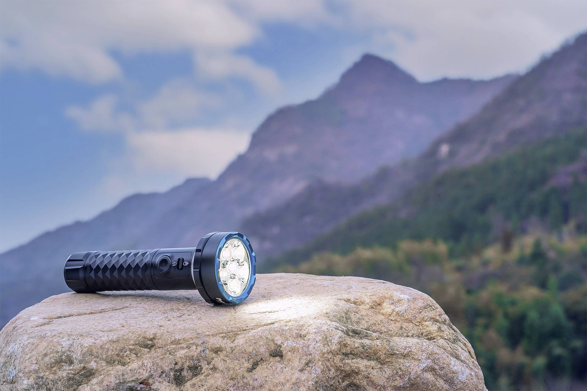 Une lampe de poche noire brille sur un grand rocher, avec en arrière-plan des montagnes boisées et un ciel nuageux.