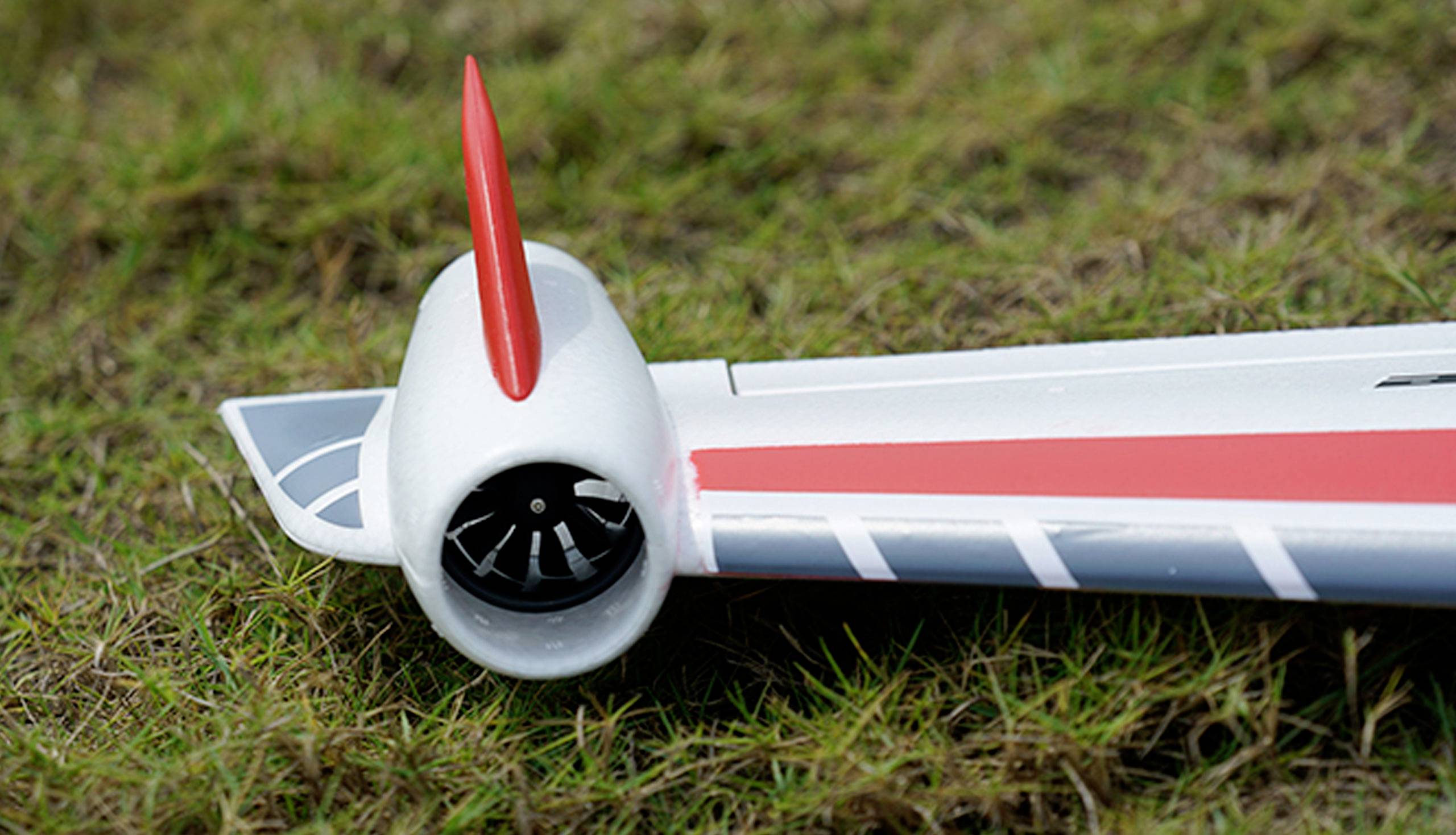 Gros plan sur l'aile d'un avion miniature avec un empennage rouge et une turbine, posée sur un sol herbeux.