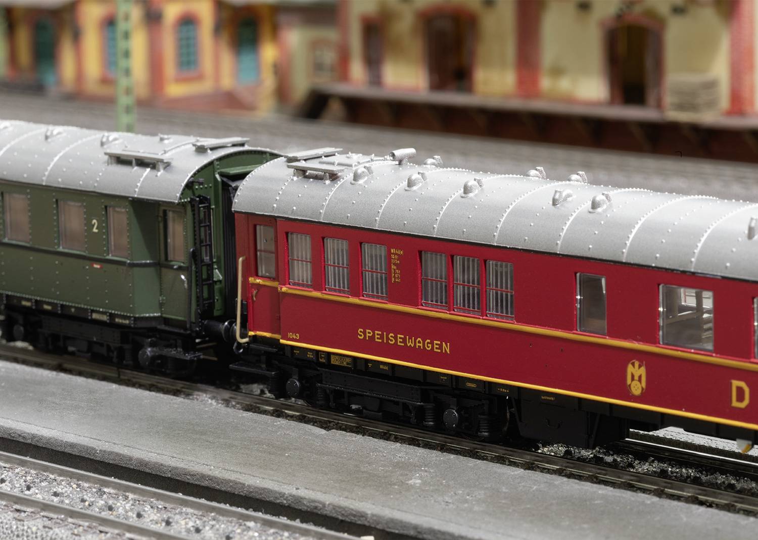 Un train miniature avec une voiture-restaurant rouge et une voiture de voyageurs verte est placé sur une voie devant un bâtiment de gare historique.