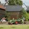 Un paysage miniature représente une ferme avec deux tracteurs verts, un agriculteur, une femme en costume traditionnel et une vache sur un chemin de gravier.
