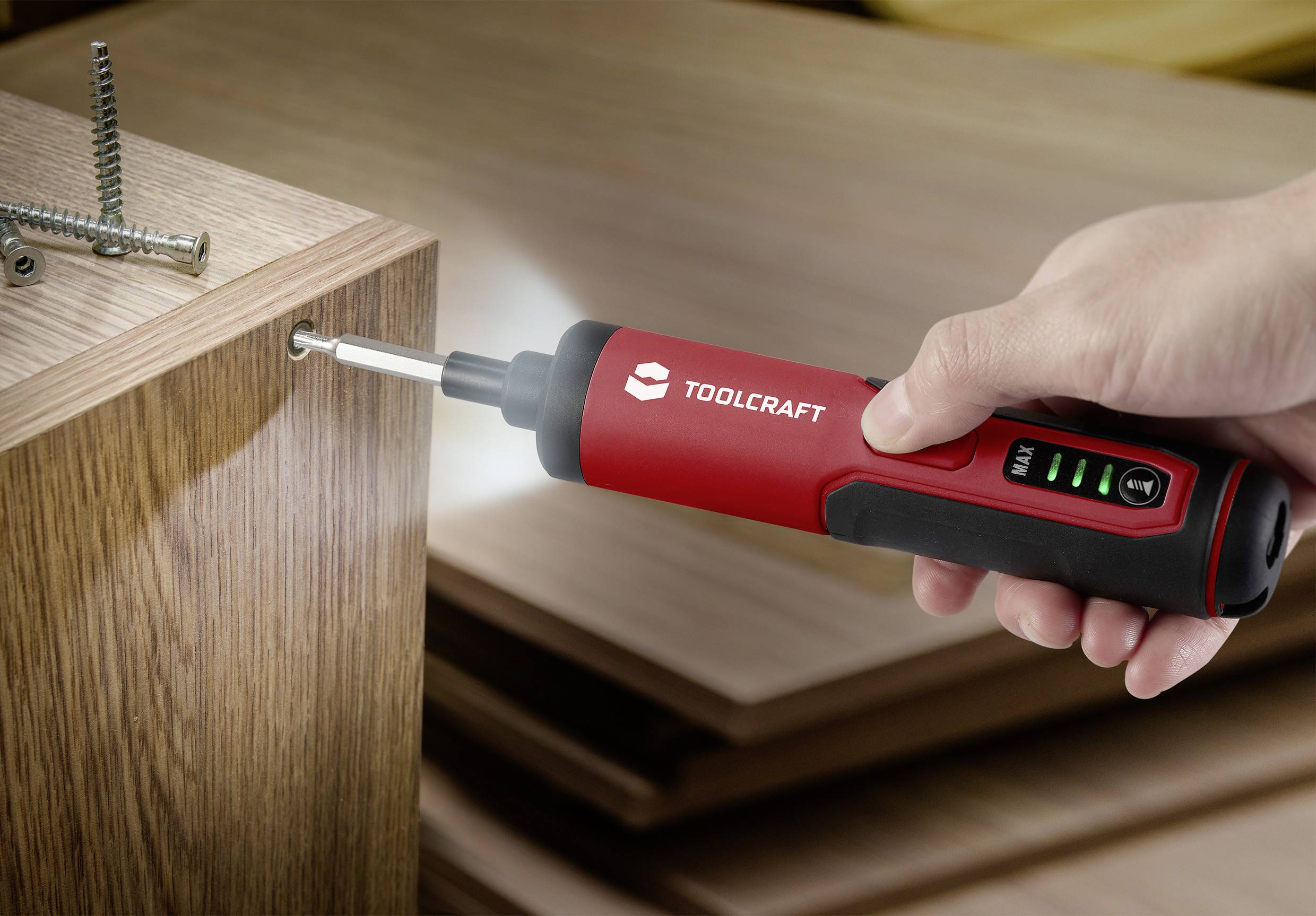 Une main tient un tournevis électrique rouge qui visse une vis dans une planche en bois. Trois vis sont fixées dans la planche.