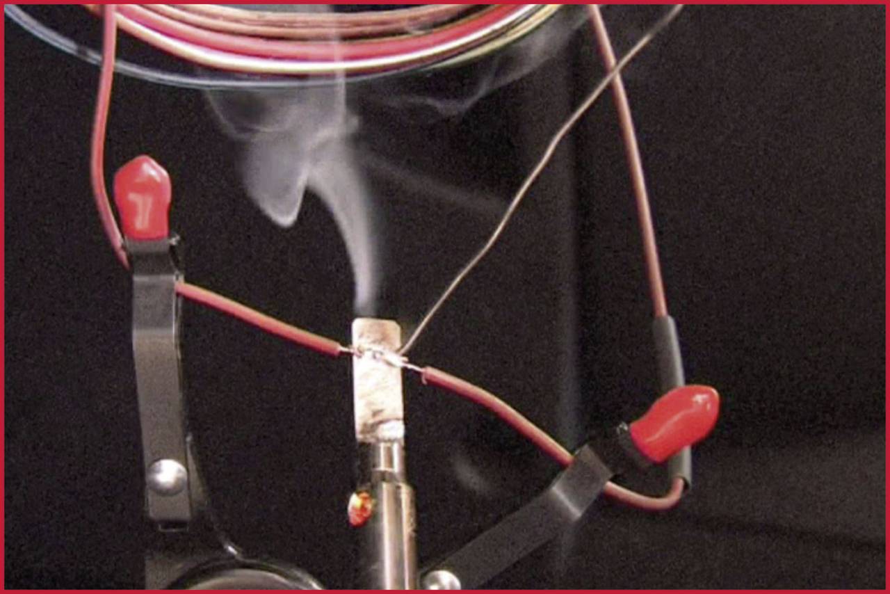 De la fumée s'élève d'un fil chauffé maintenu par des pinces crocodiles. Expérience électrique démontrant le passage du courant.