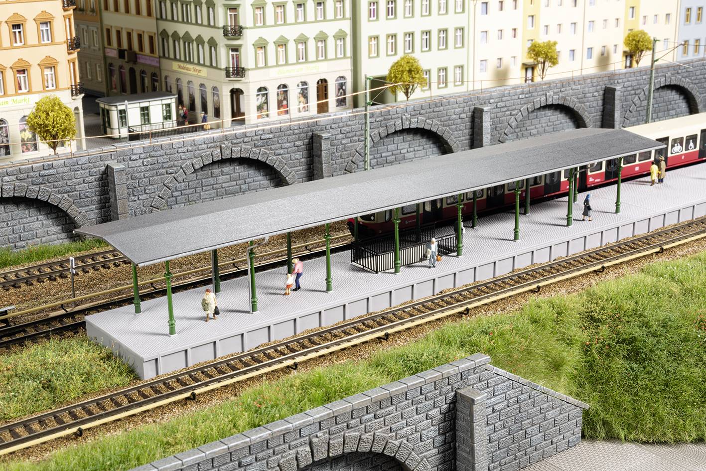 Scène de train miniature avec une gare, un train, des voies ferrées et des personnages attendant sur le quai. L'arrière-plan montre des immeubles urbains et un pont.