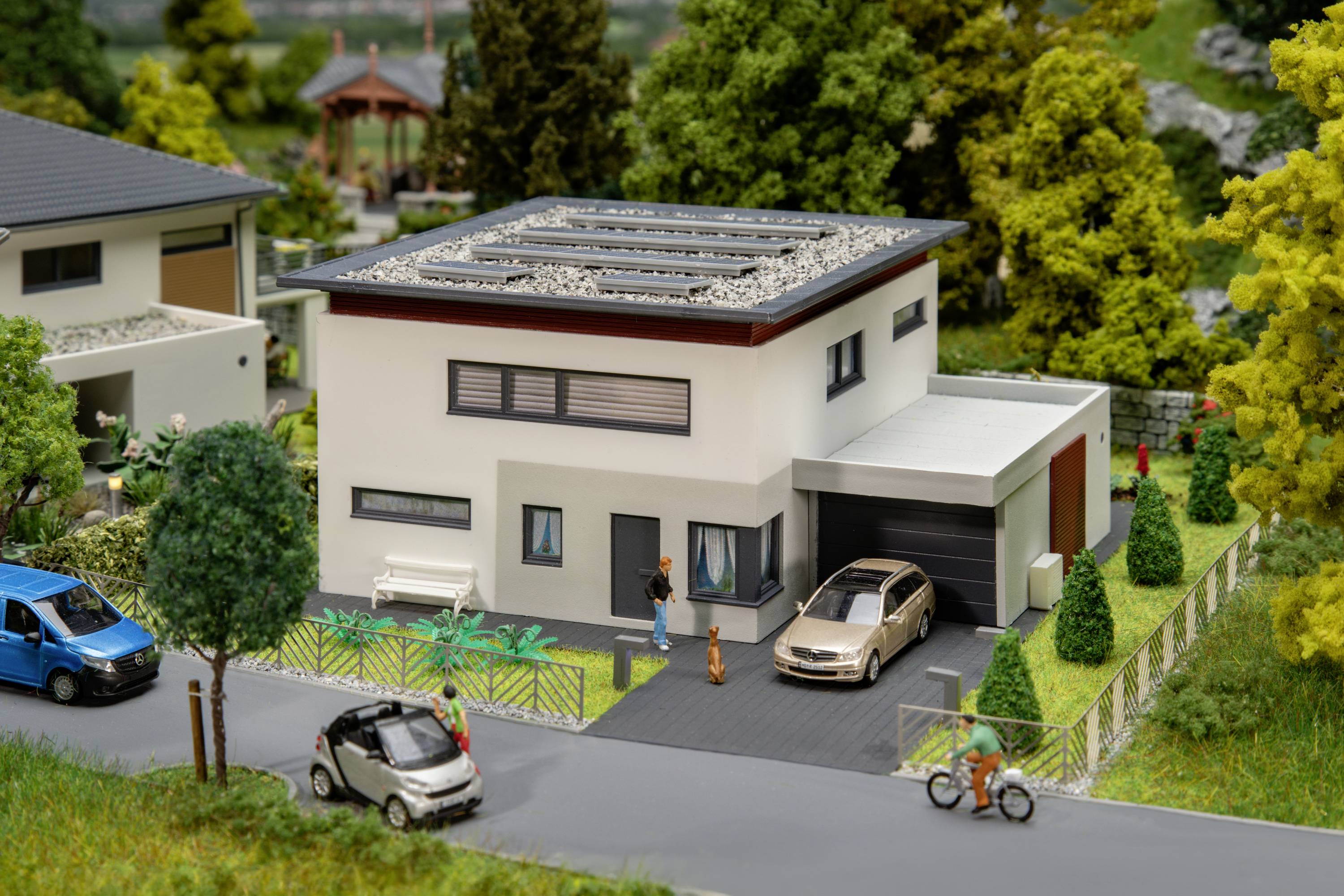 Une maison individuelle moderne avec un toit plat et des panneaux solaires, entourée d'arbres. Une famille se tient devant la maison, une voiture est garée dans l'allée.