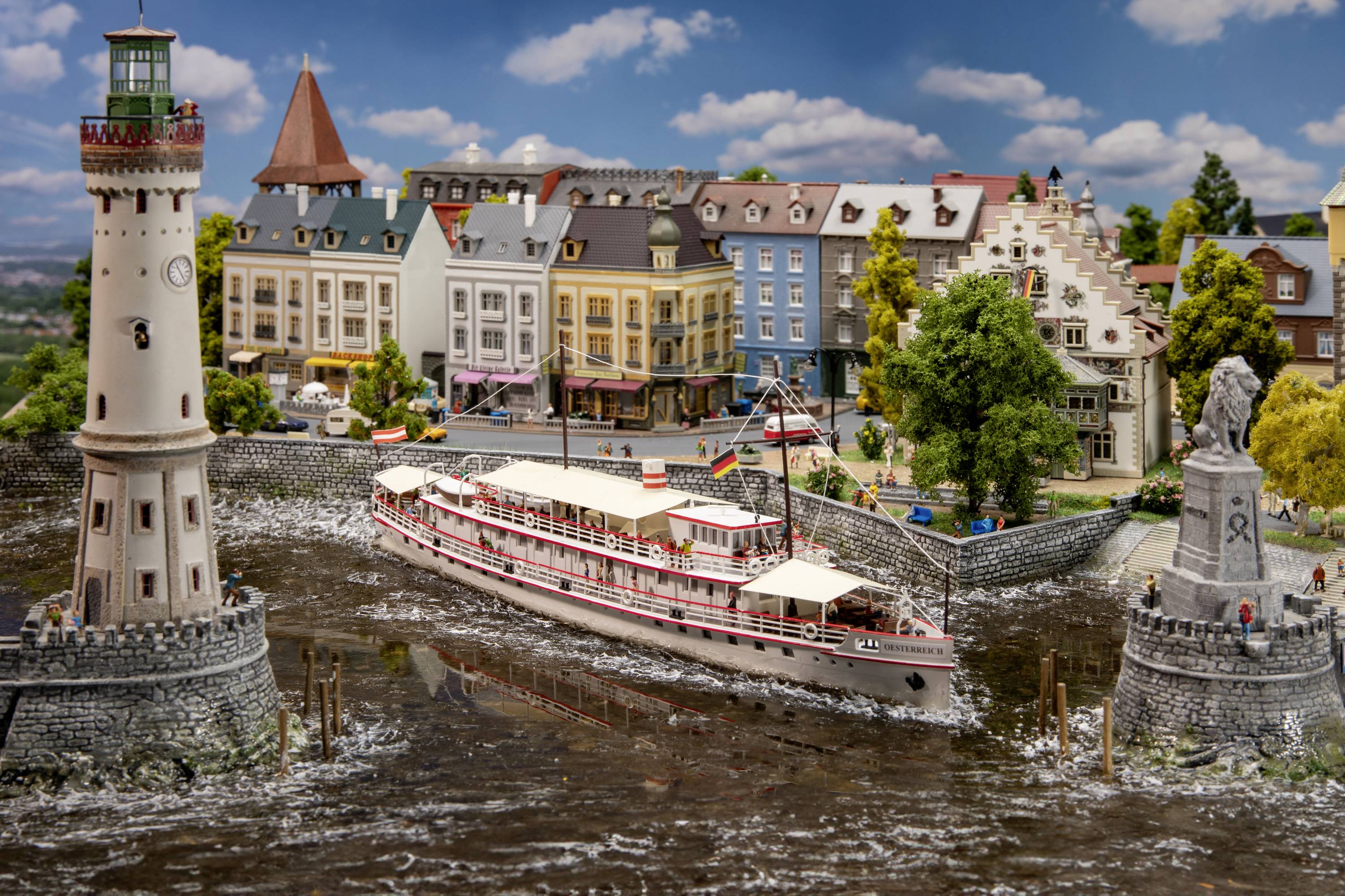Un bateau-modèle navigue à travers un canal miniature, flanqué d'un phare et de bâtiments en arrière-plan.