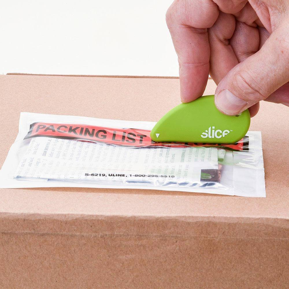 Une main utilise un petit coupe-ruban vert pour ouvrir un ruban adhésif sur un carton le long d'une liste de colisage rouge.
