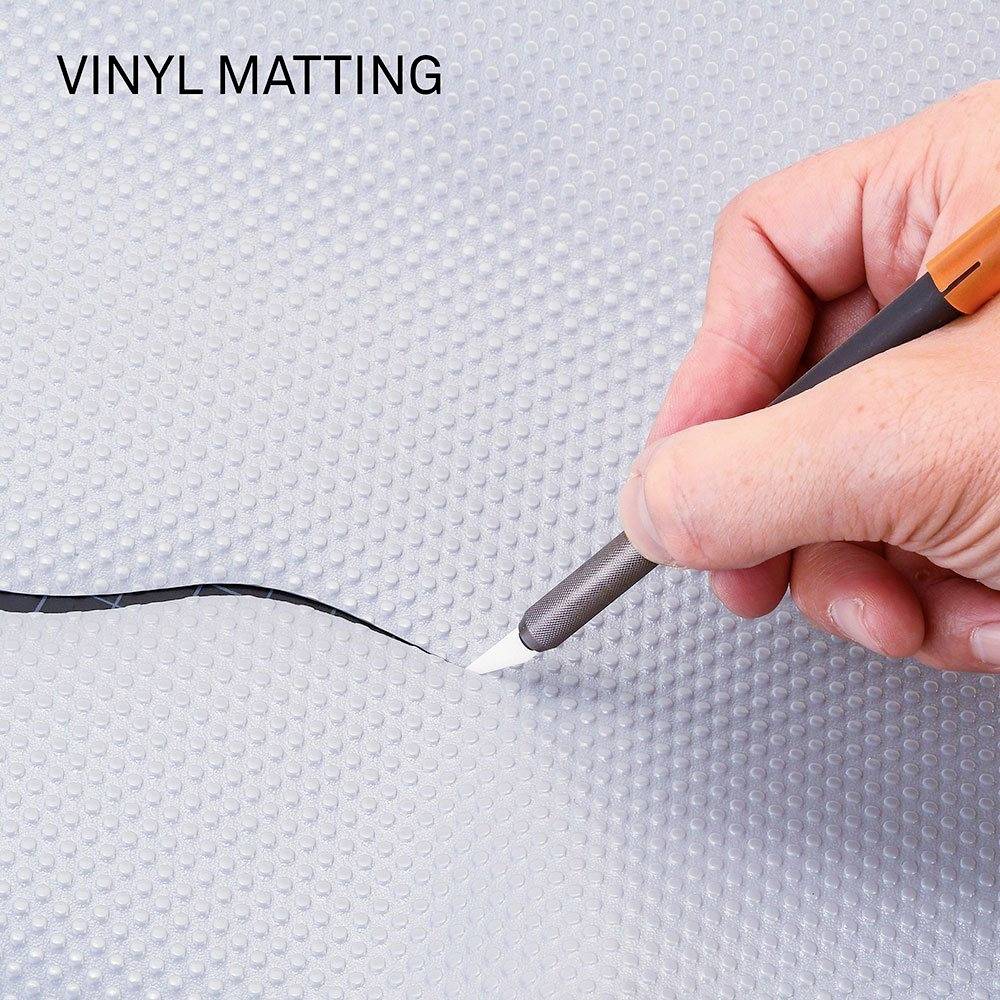 Une main coupe une bande noire dans un tapis de vinyle gris à l'aide d'un couteau.