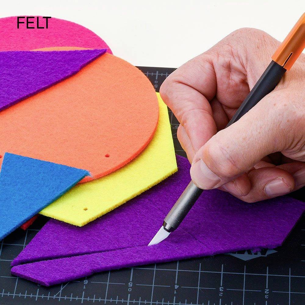 Une main coupe du feutre violet avec un cutter sur une planche de coupe avec règle. Des morceaux de feutre colorés sont visibles en arrière-plan.