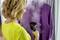 Une femme utilise un fer à repasser à vapeur pour lisser une chemise violette. Elle porte un haut jaune. Des rideaux sont visibles en arrière-plan.