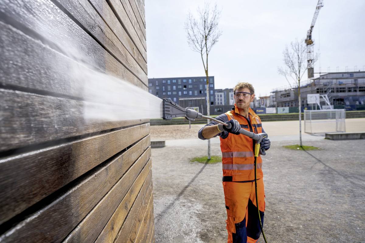 Une personne portant un équipement de sécurité orange utilise un nettoyeur haute pression sur un mur en bois sur un chantier de construction, avec des bâtiments et une grue visibles en arrière-plan.