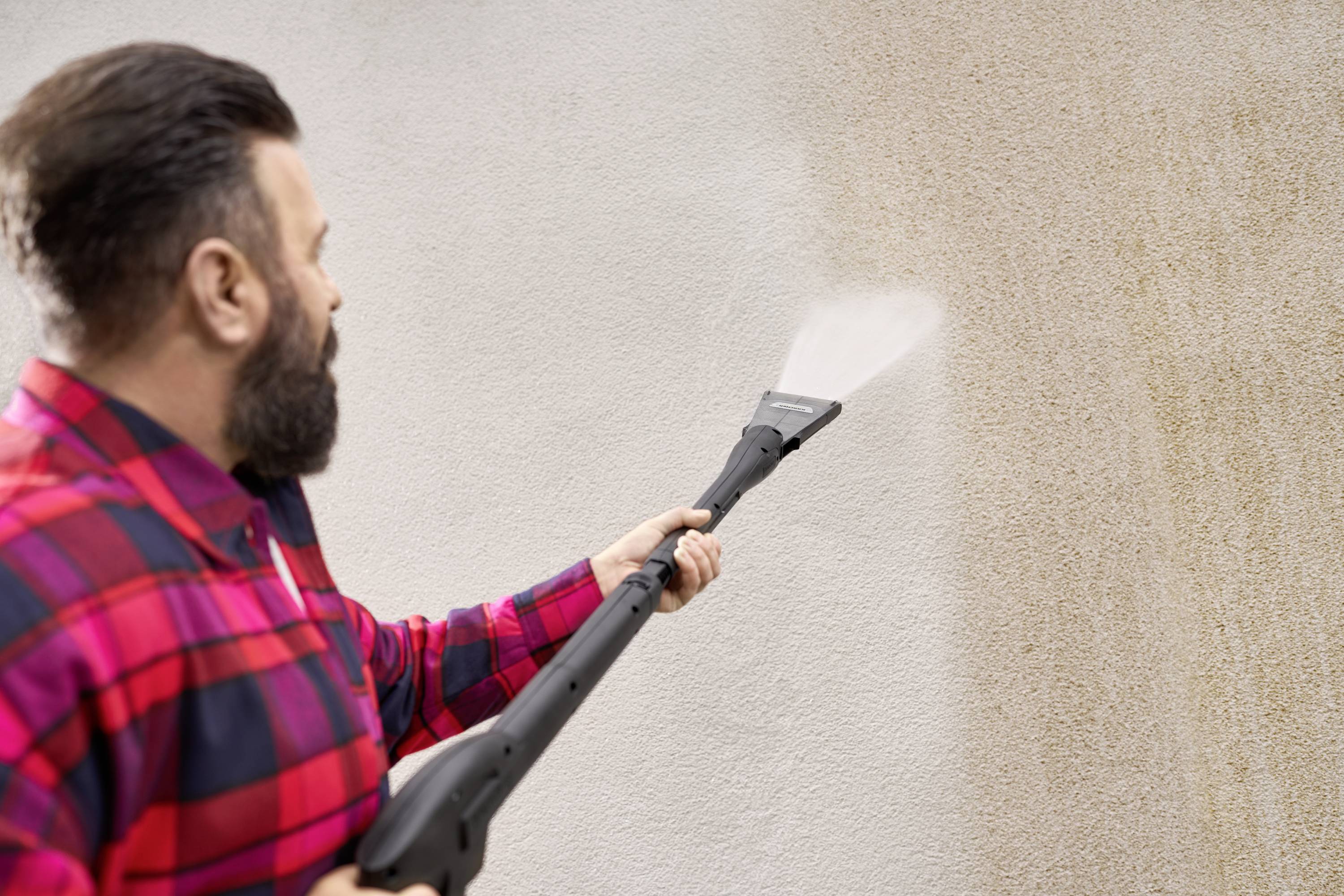 Une personne portant une chemise à carreaux nettoie un mur sale à l'aide d'un nettoyeur haute pression. Le mur devient visiblement plus propre.