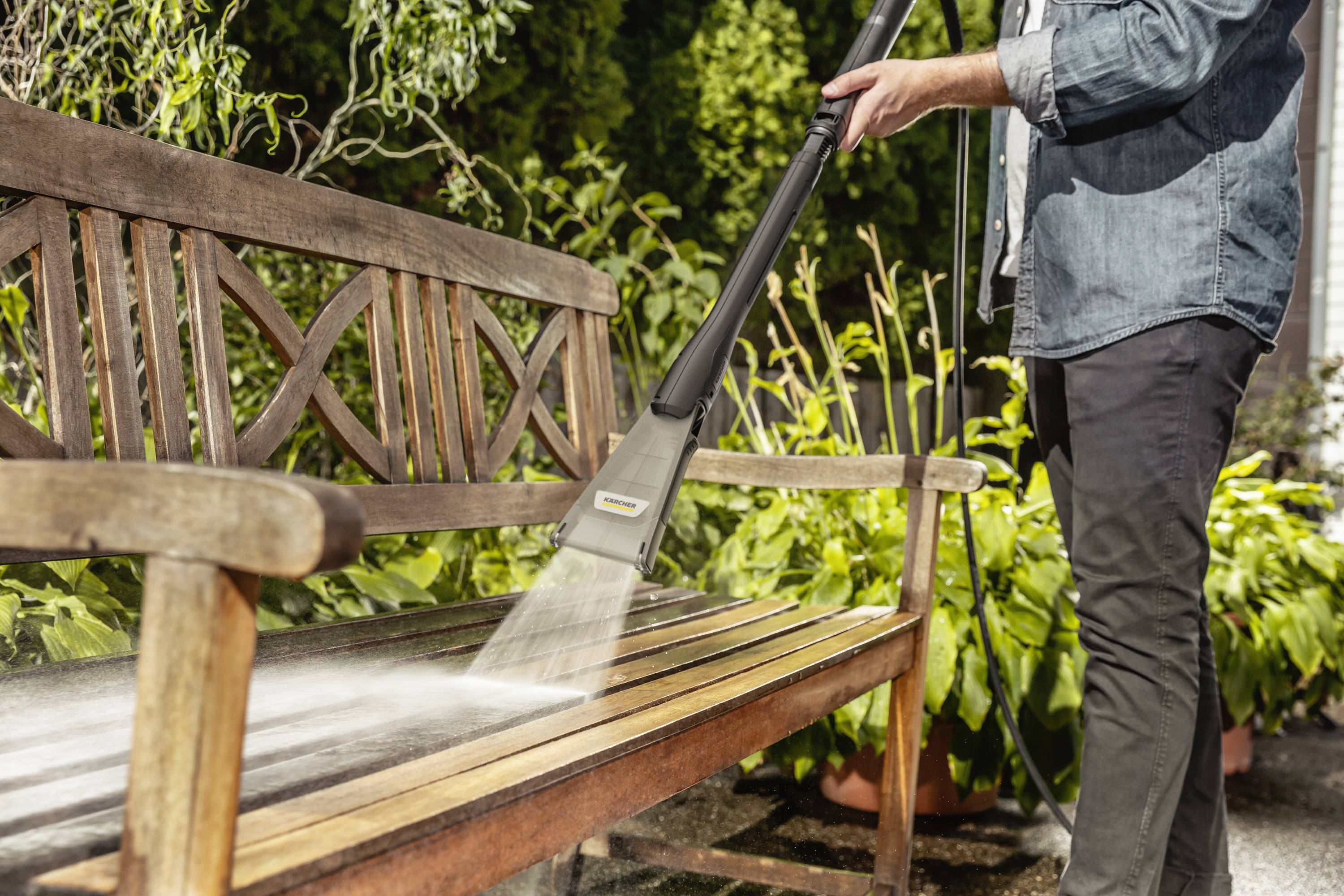 Une personne nettoie un banc en bois dans le jardin à l'aide d'un nettoyeur haute pression. Des plantes et des arbres sont visibles en arrière-plan.
