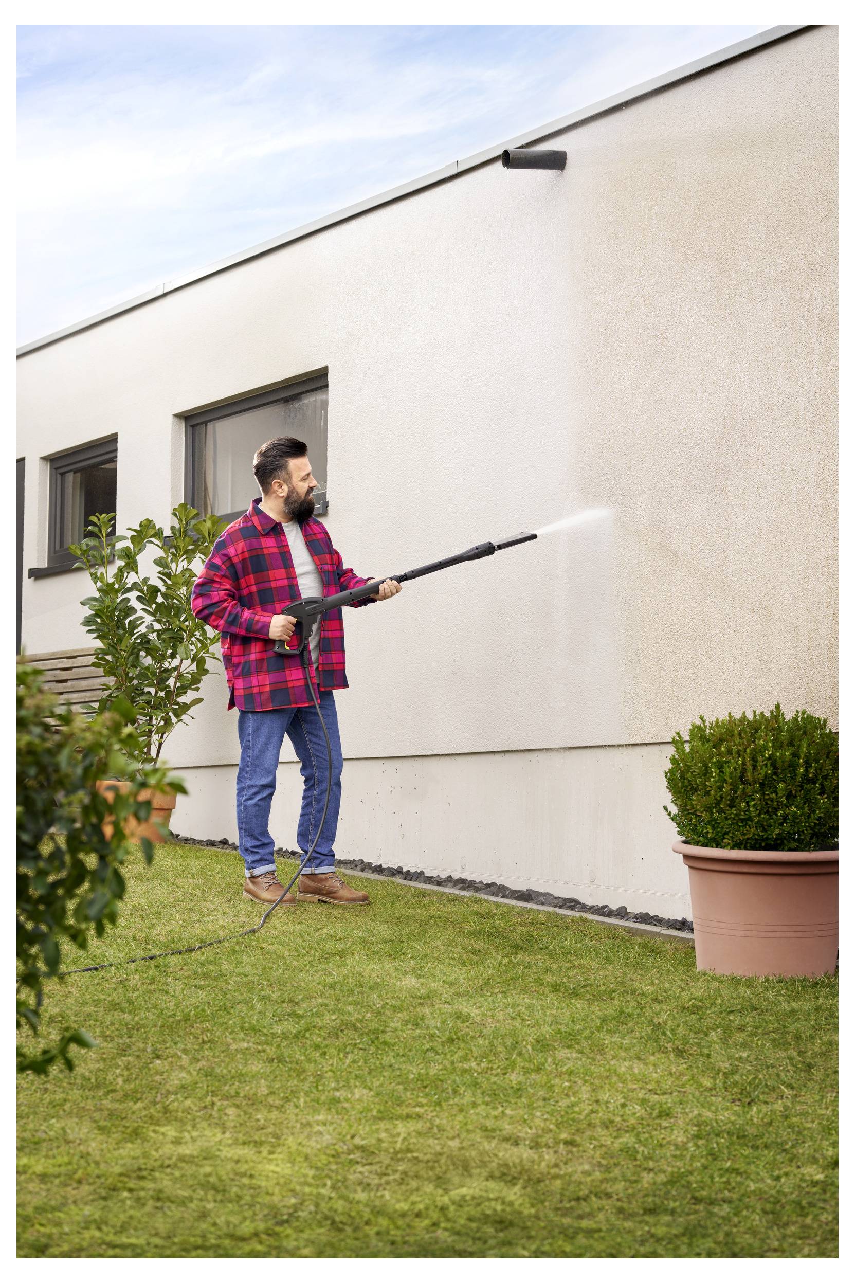 Une personne portant une chemise de flanelle rouge utilise un nettoyeur haute pression pour nettoyer le mur extérieur d'une maison dans un jardin avec de l'herbe verte et des plantes en pot.