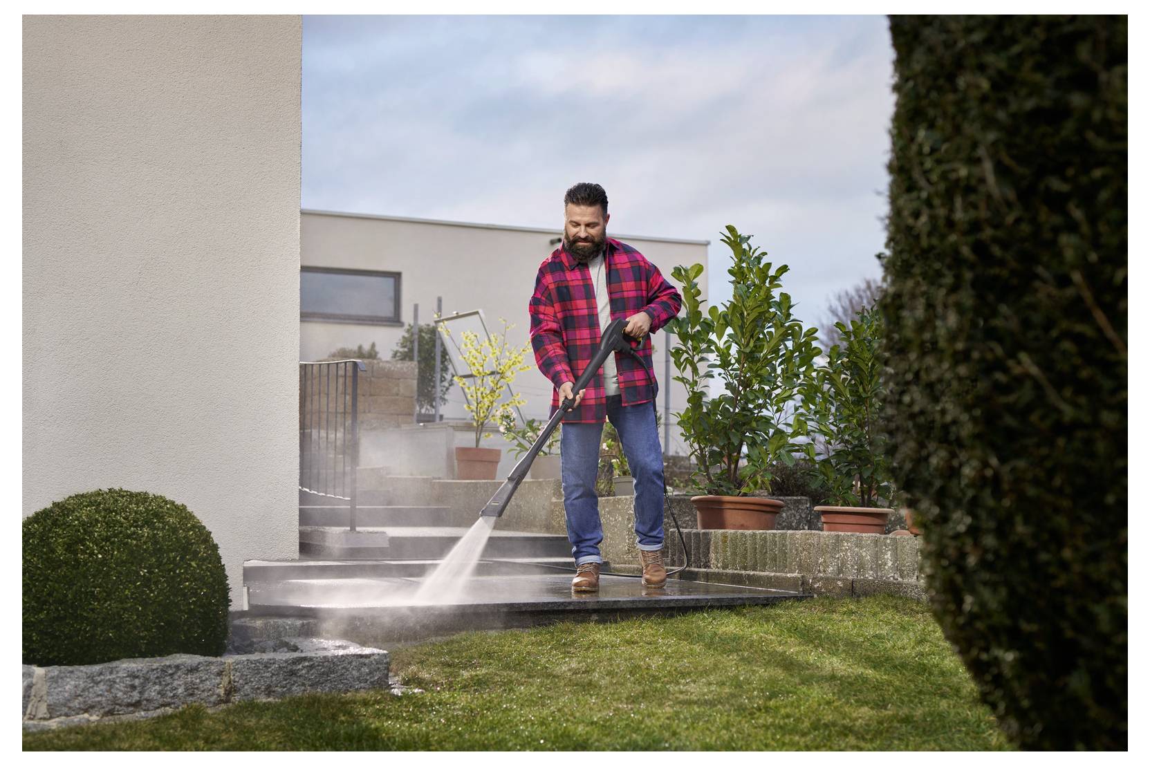 Homme portant une chemise de flanelle rouge utilisant un nettoyeur haute pression sur une terrasse en pierre ; des plantes en pot et des haies taillées sont visibles en arrière-plan ; scène de nettoyage extérieur.