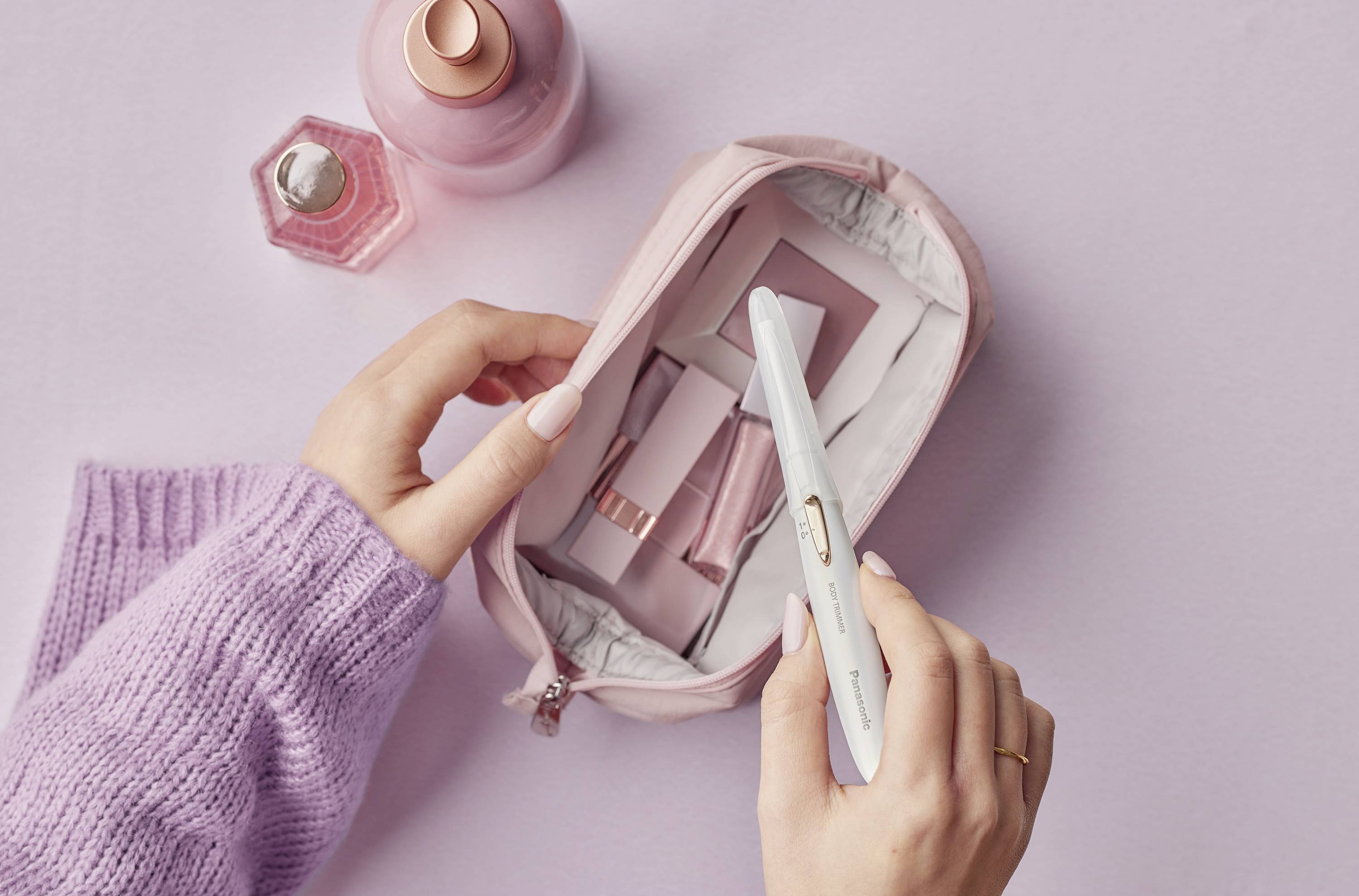 Une femme range des articles de cosmétique dans une trousse de toilette rose, sur un fond lilas. En haut à gauche se trouvent deux flacons de parfum.