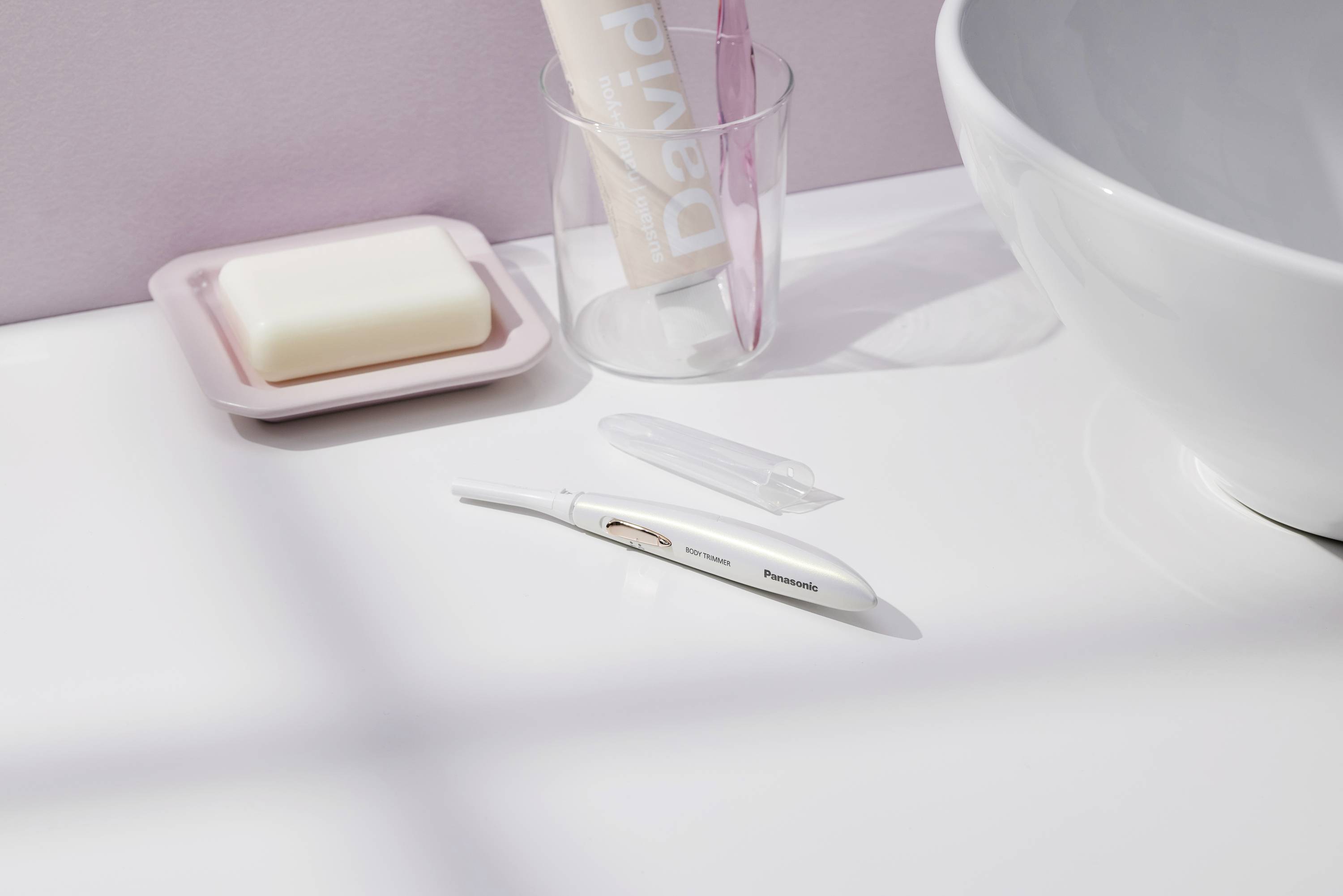 Brosse à dents électrique sur une surface blanche de salle de bain, à côté d'un savon dans un porte-savon et un verre transparent contenant du dentifrice et une brosse à dents rose.