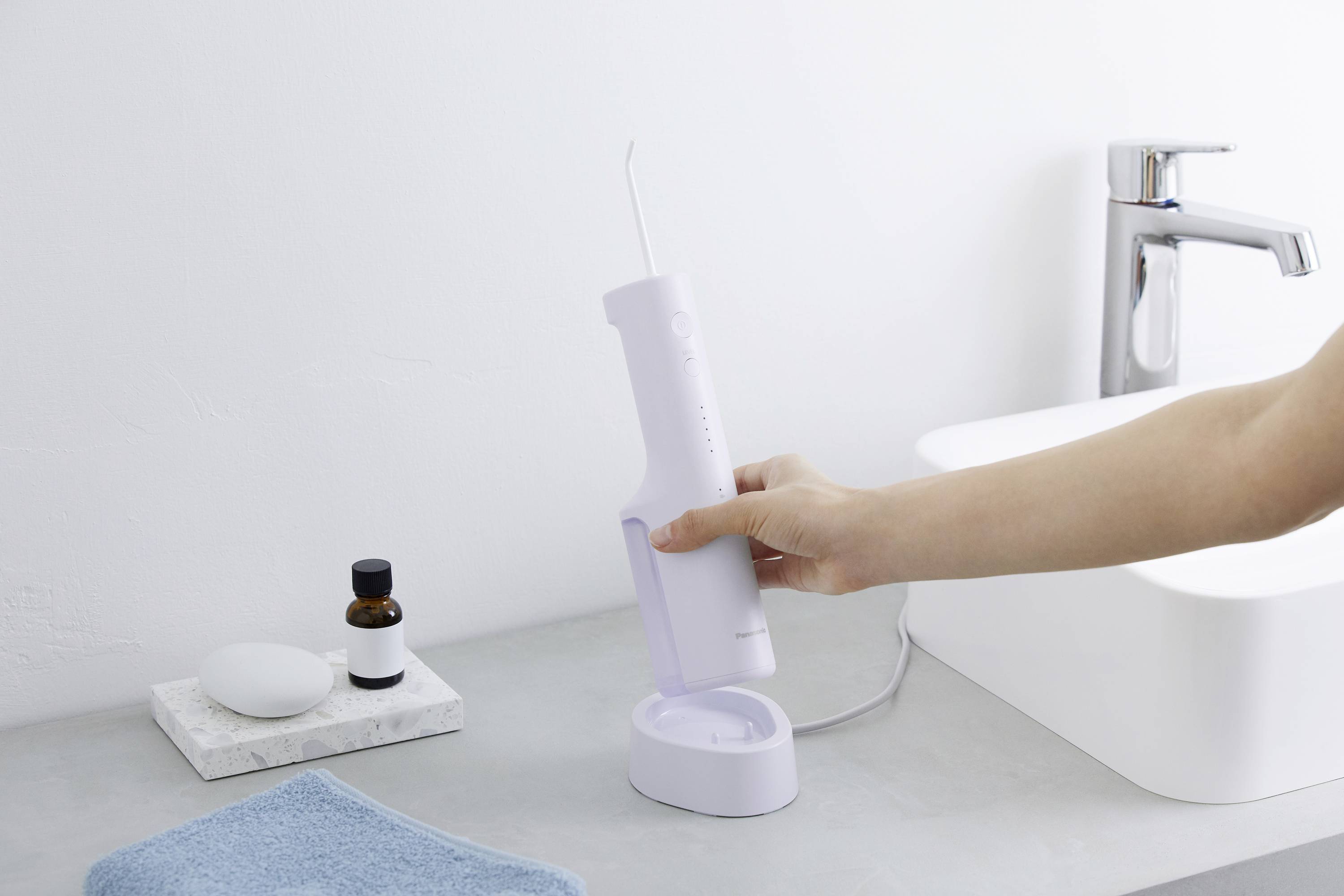 Une personne tient un irrigateur buccal blanc devant un lavabo. À côté se trouvent une serviette, un distributeur de savon et une bouteille.