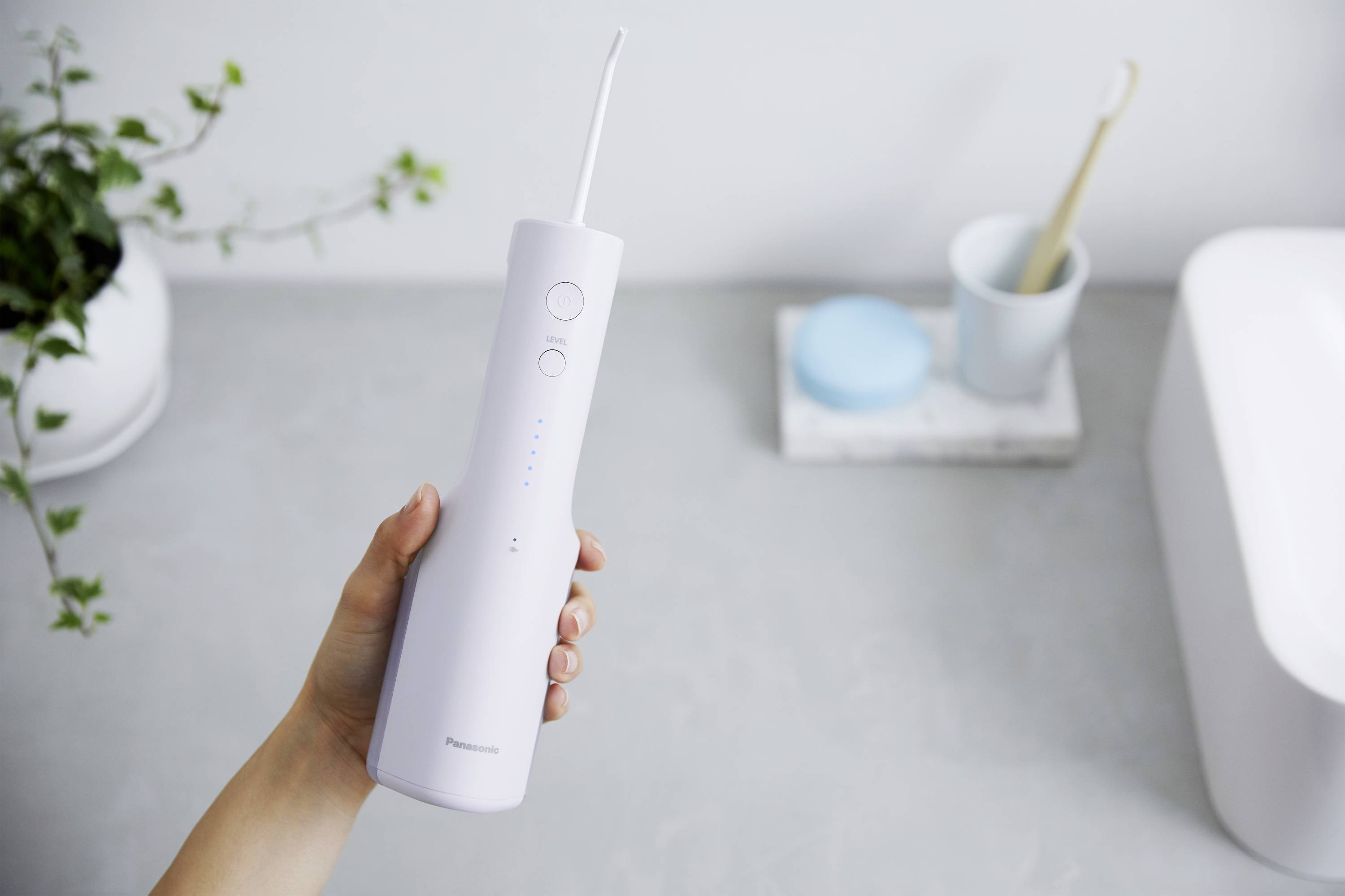 Une main tient un irrigateur buccal électrique au-dessus d'un meuble de salle de bain gris ; des plantes et une brosse à dents sont visibles en arrière-plan.