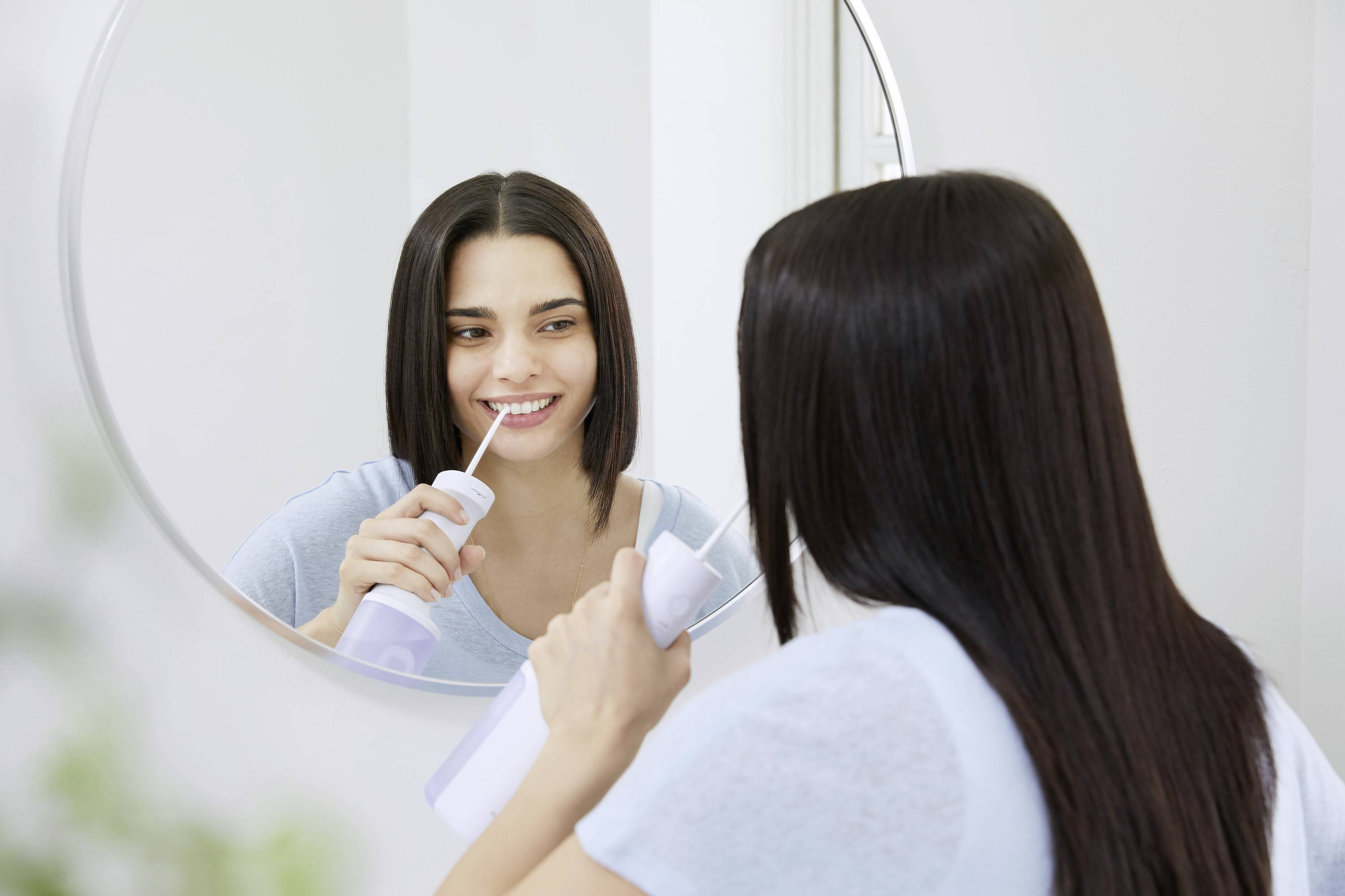 Une femme utilise un jet dentaire devant un miroir. Elle sourit, portant un t-shirt bleu clair. Environnement de salle de bain lumineux et minimaliste.