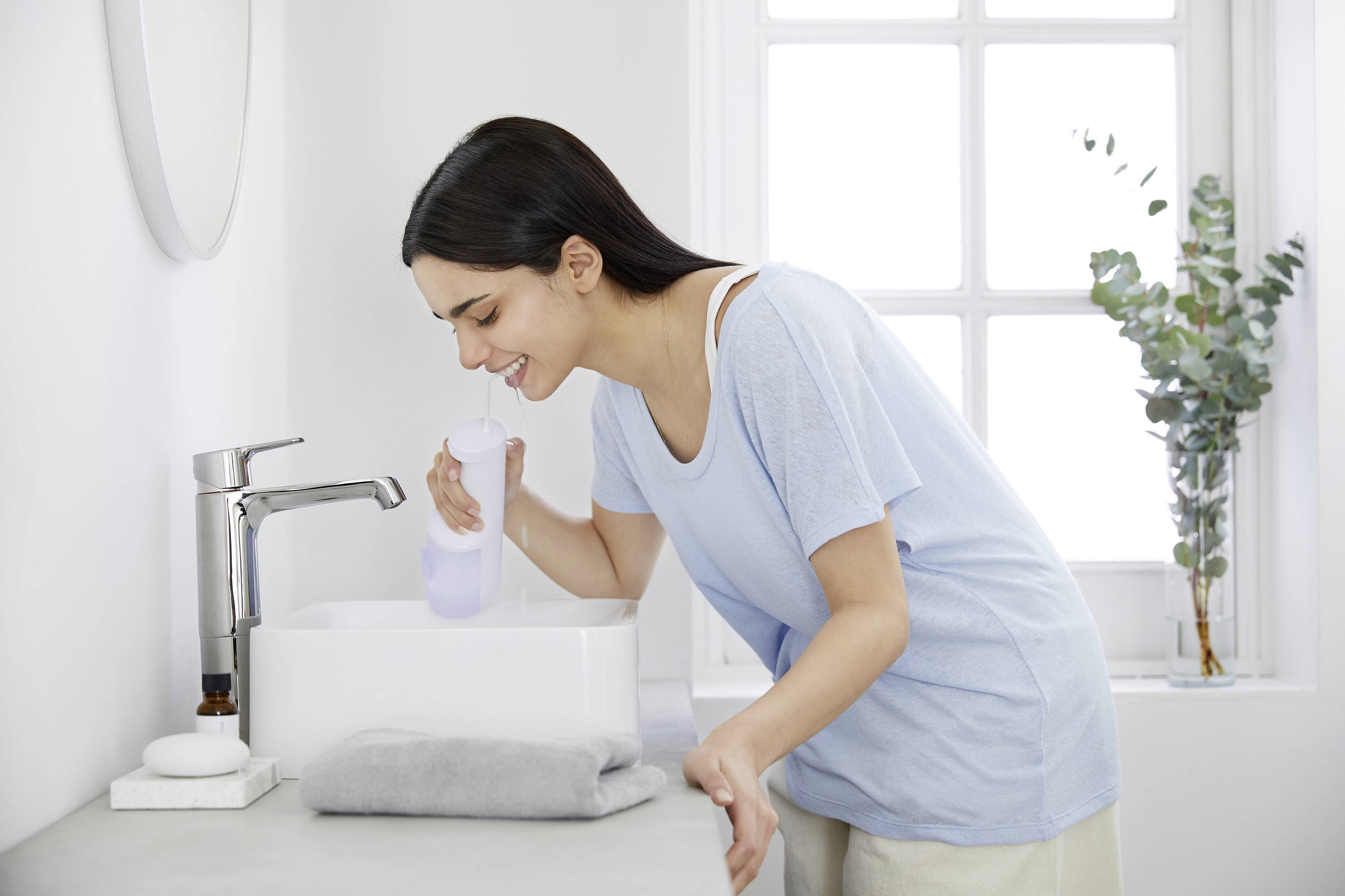 Une femme portant un t-shirt bleu clair utilise un irrigateur buccal au-dessus d'un lavabo dans une salle de bain moderne avec une grande fenêtre et des plantes.