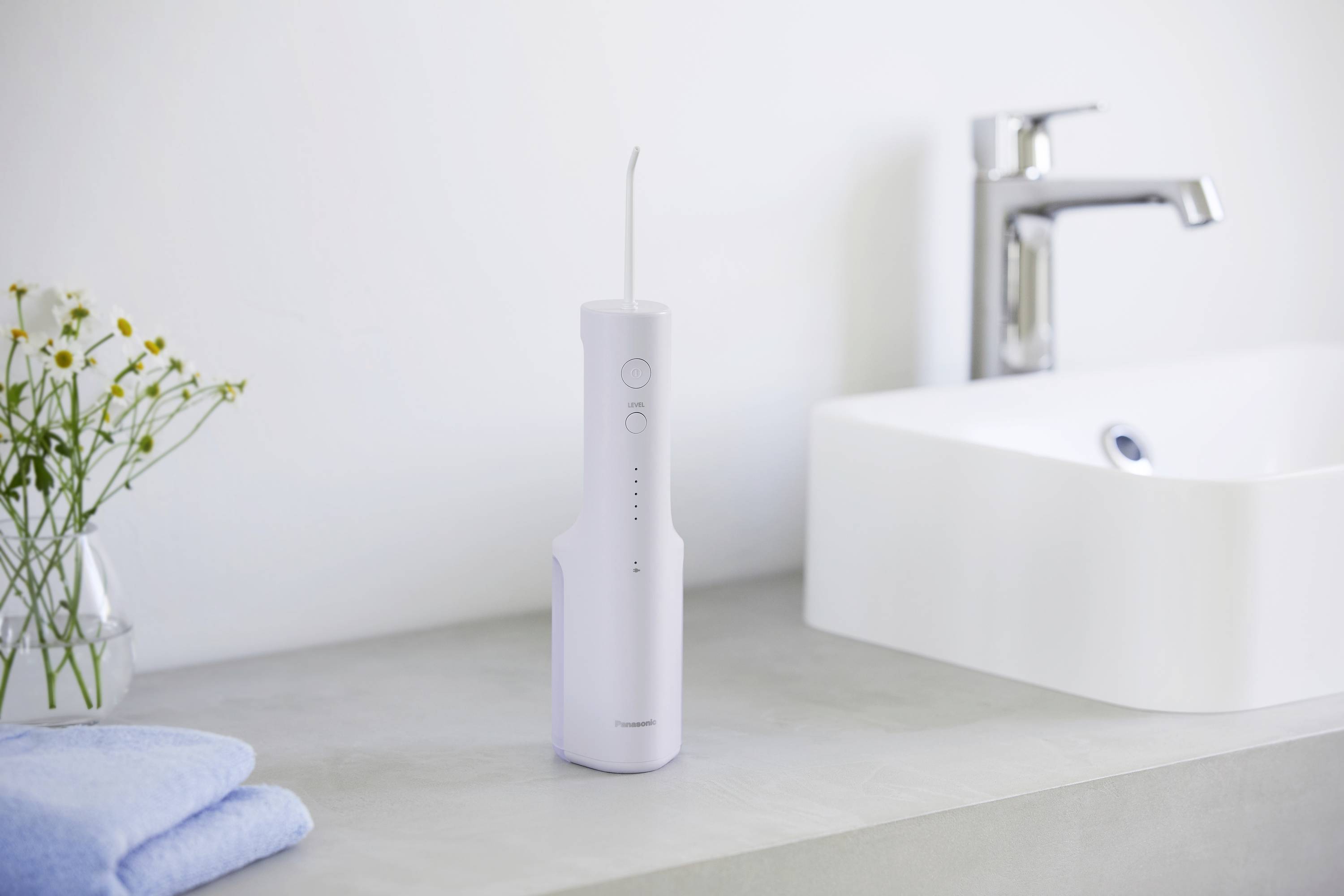 Douche buccale électrique sur le lavabo à côté d'un robinet, avec une serviette et des fleurs en arrière-plan.