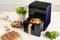 Un airfryer noir avec une poignée dorée est posé sur un plan de travail de cuisine. À l'intérieur, un poulet rôti accompagné de légumes. À côté, du pain, des frites et des herbes.