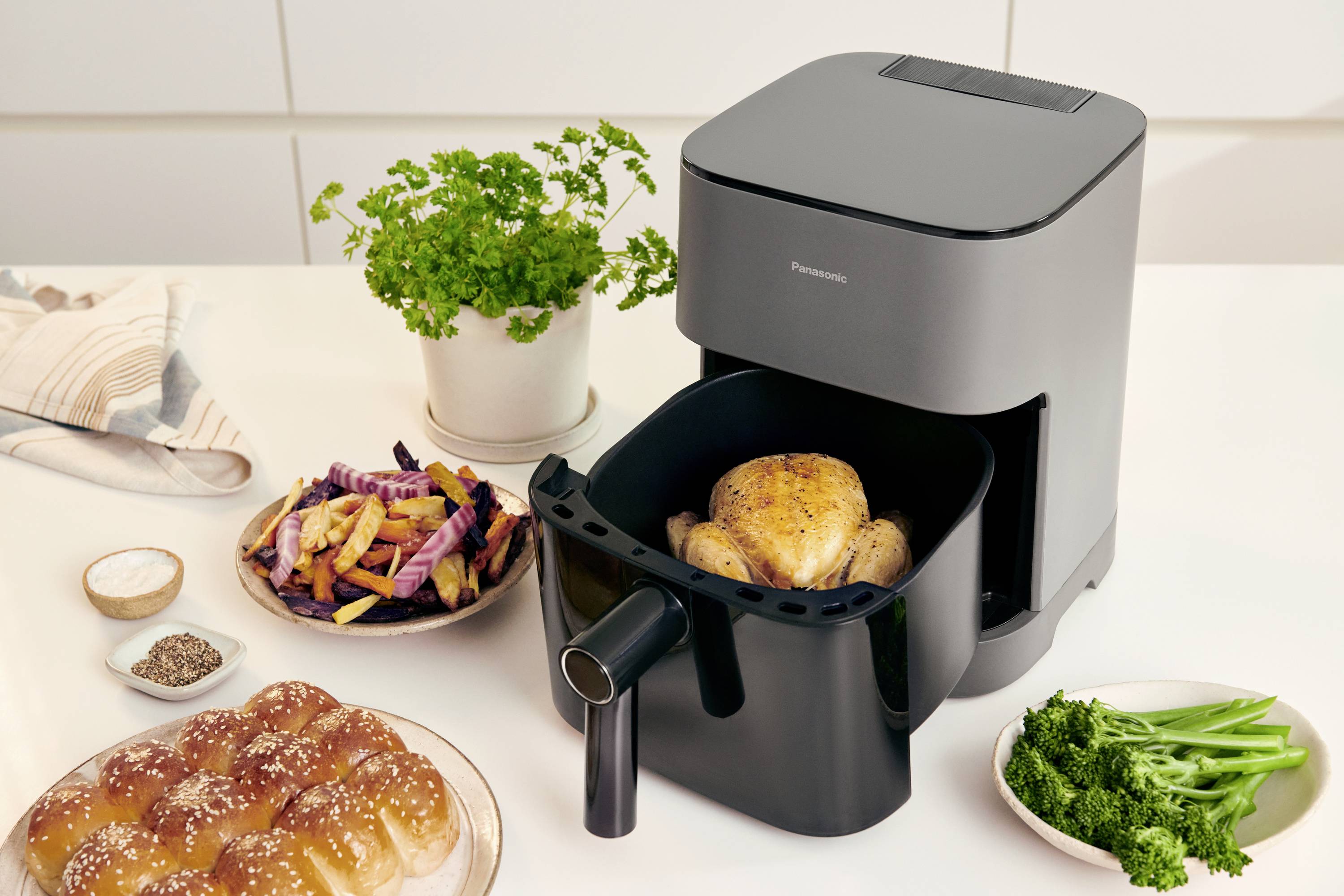 Une friteuse à air chaud grise avec un poulet rôti. À côté, un plateau avec des frites colorées, des petits pains et du persil frais est posé sur une table.