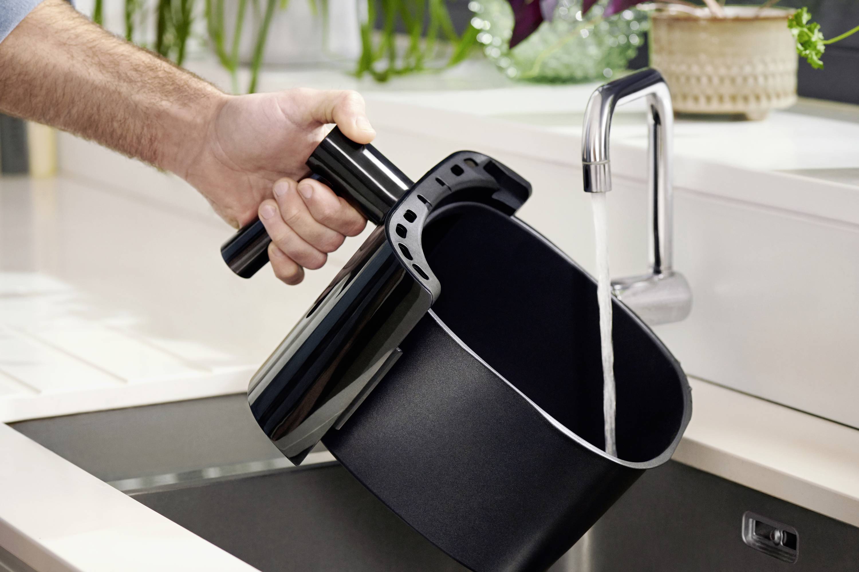 Une main tient un panier de friteuse à air qui est rincé sous un robinet.