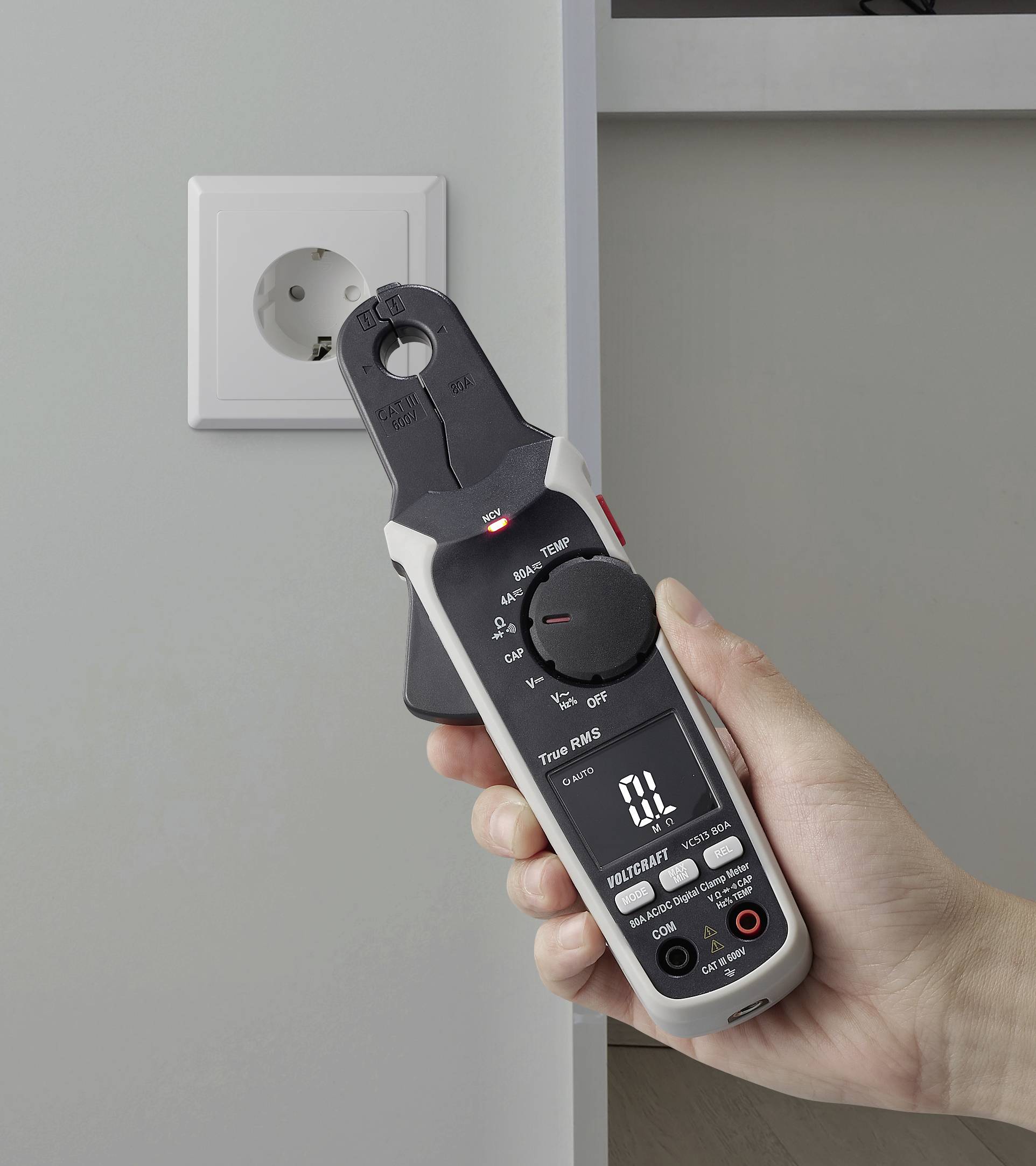 Une main tient un multimètre numérique mesurant une prise de courant. L'affichage du multimètre montre des relevés, indiquant qu'il est utilisé pour un test électrique.