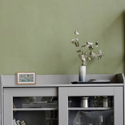 Un meuble gris devant un mur vert, sur lequel se trouvent un vase avec des fleurs séchées et un calendrier numérique.