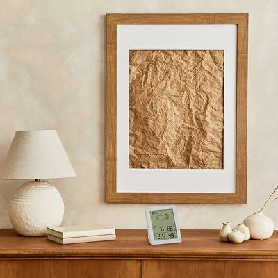 Une table en bois avec une lampe, deux livres, un thermomètre numérique et de petites céramiques. Un tableau accroché au mur avec du papier brun.