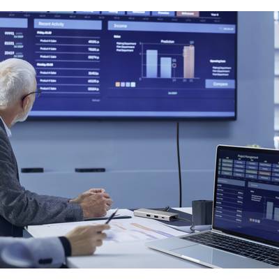 Un homme âgé est assis à une table de réunion et examine des diagrammes et des données sur un grand écran, qui affichent des données de performance.