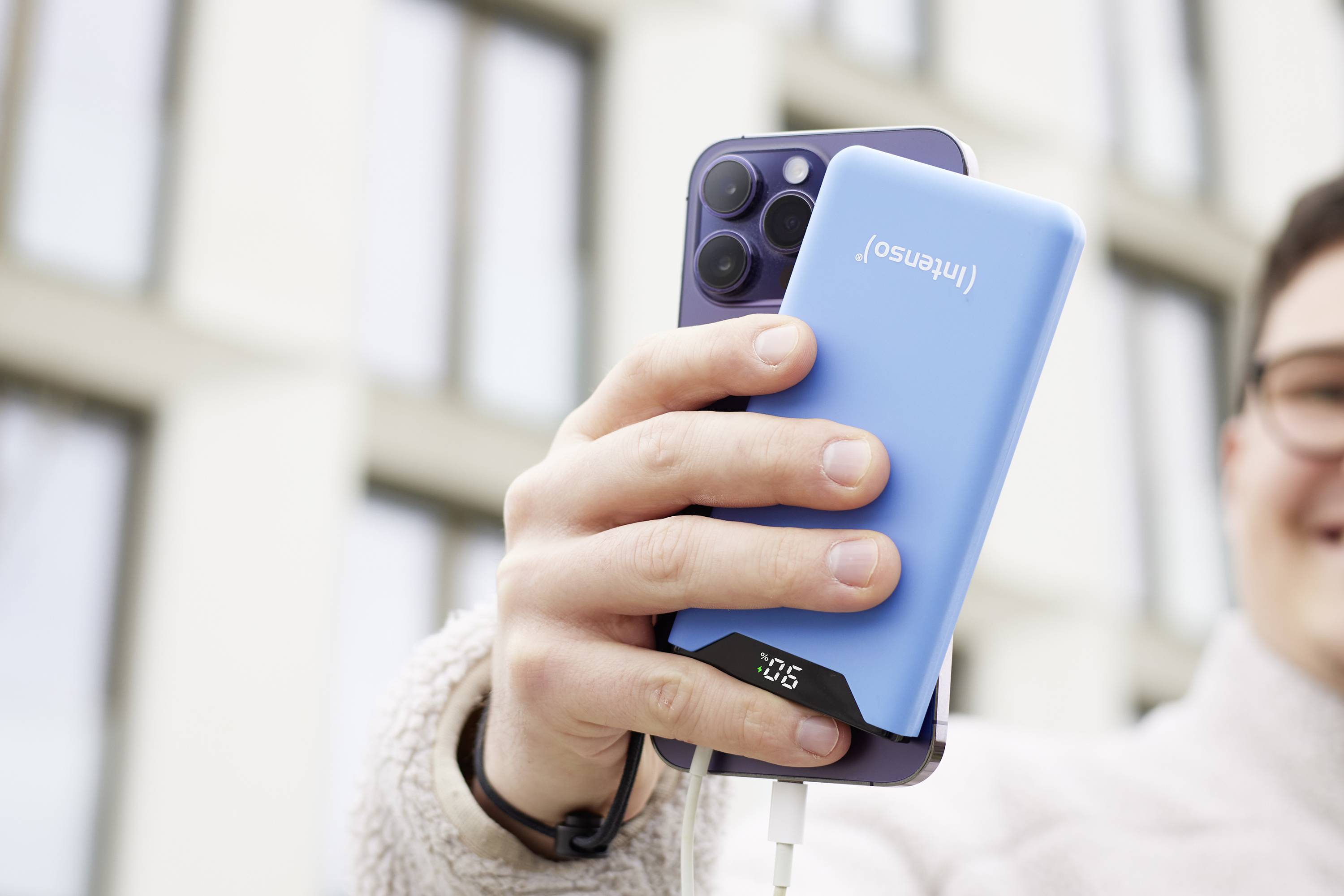 Une personne tient un smartphone et une batterie externe bleue avec un câble branché dans sa main. Des bâtiments sont visibles en arrière-plan.