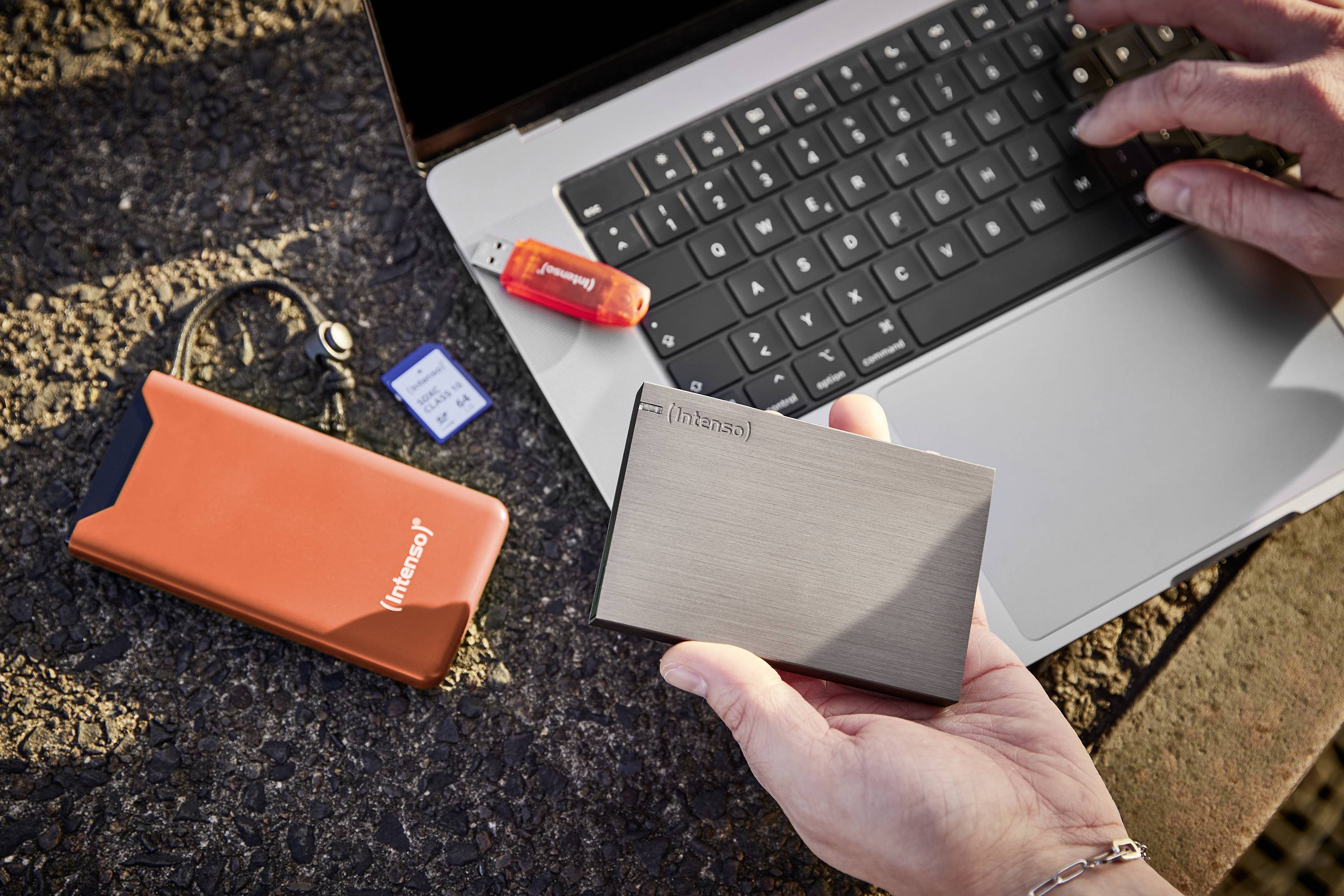 Une personne tient un disque dur externe devant un ordinateur portable. Sur une table sont posés une clé USB, une carte SD et une housse orange.