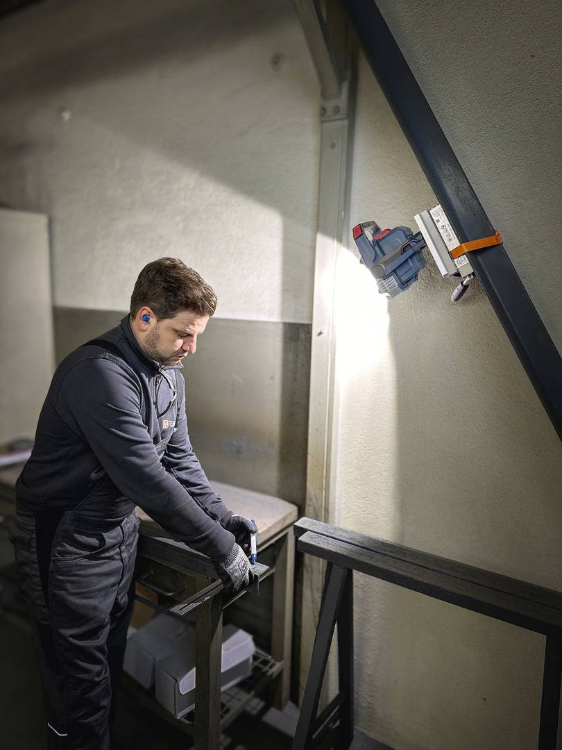 Un homme en combinaison de travail coupe du métal avec une scie dans un atelier. L'environnement est industriel et bien éclairé.