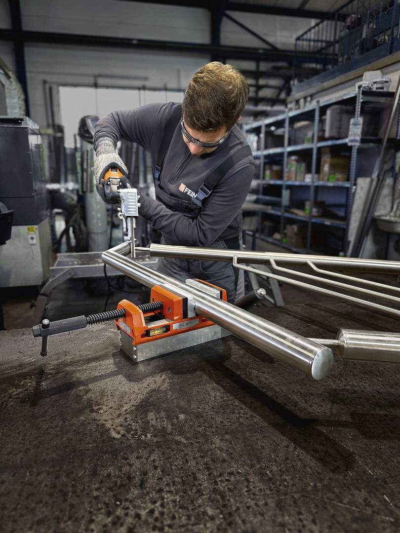 Un ouvrier en tenue de protection fore un élément métallique maintenu dans un étau orange, dans un environnement d'atelier.