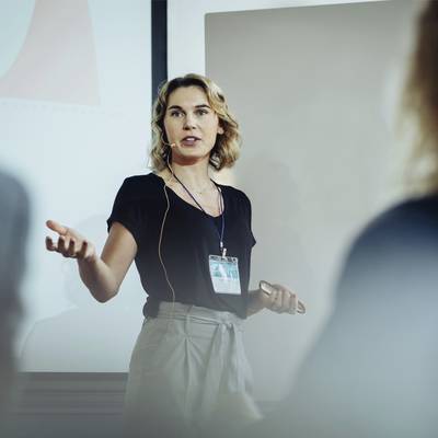 Une femme fait une présentation dans une salle de réunion, porte un casque et pointe vers un écran de projection avec des diagrammes en arrière-plan.